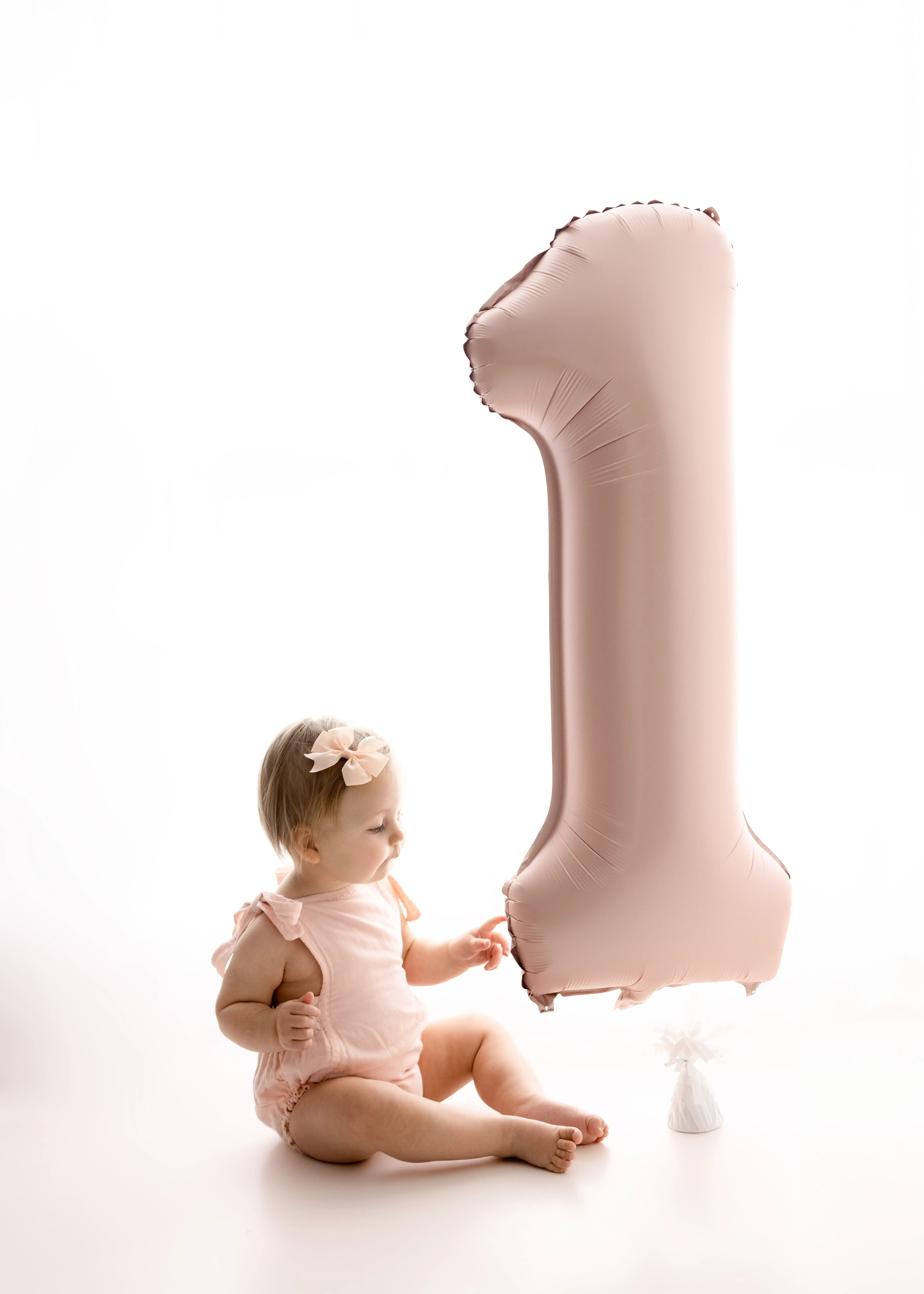 Baby girl dressed in a puffy white tutu, nibbling on a wooden spoon near a small frosted cake, celebrating her first birthday cake smash in an airy, all-white studio.