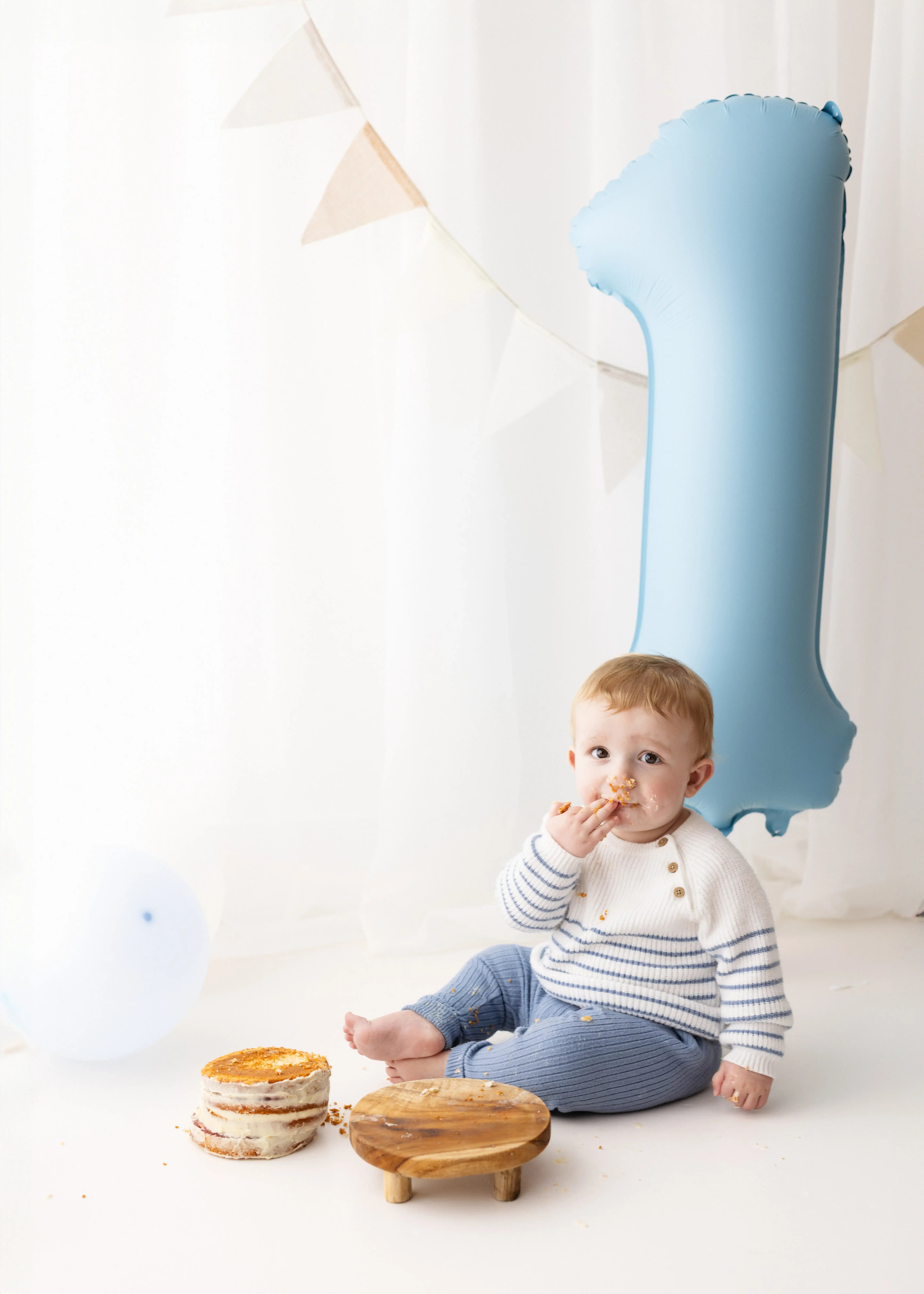 One-year-old boy wearing a white and blue striped jumper, nibbling on his first birthday cake while seated beside a large pastel-blue number one balloon in a bright, minimalist studio.