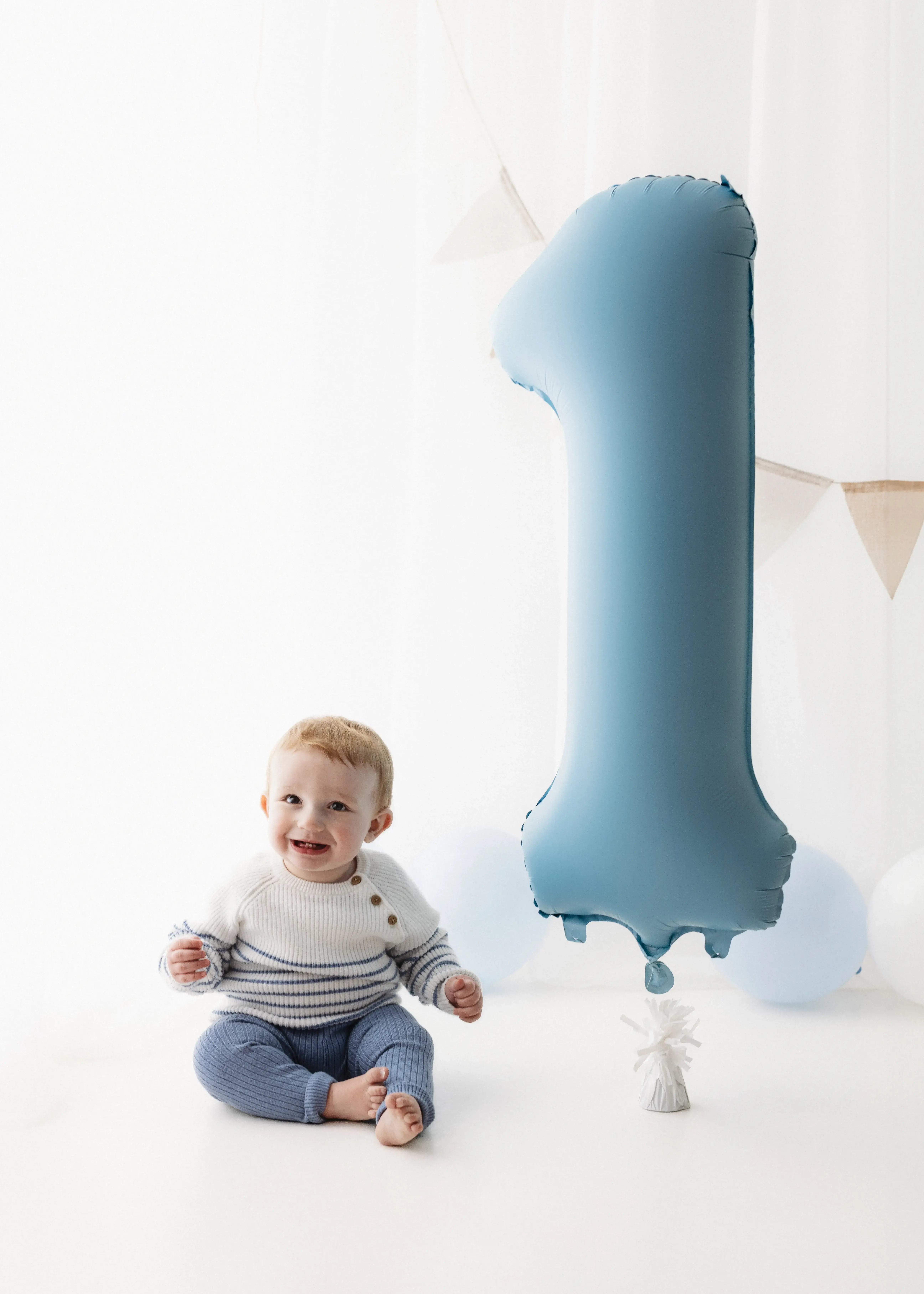 One-year-old boy sitting next to an oversized blue number one balloon, wearing a white-and-blue sweater and smiling widely in a bright, banner-adorned photography studio.