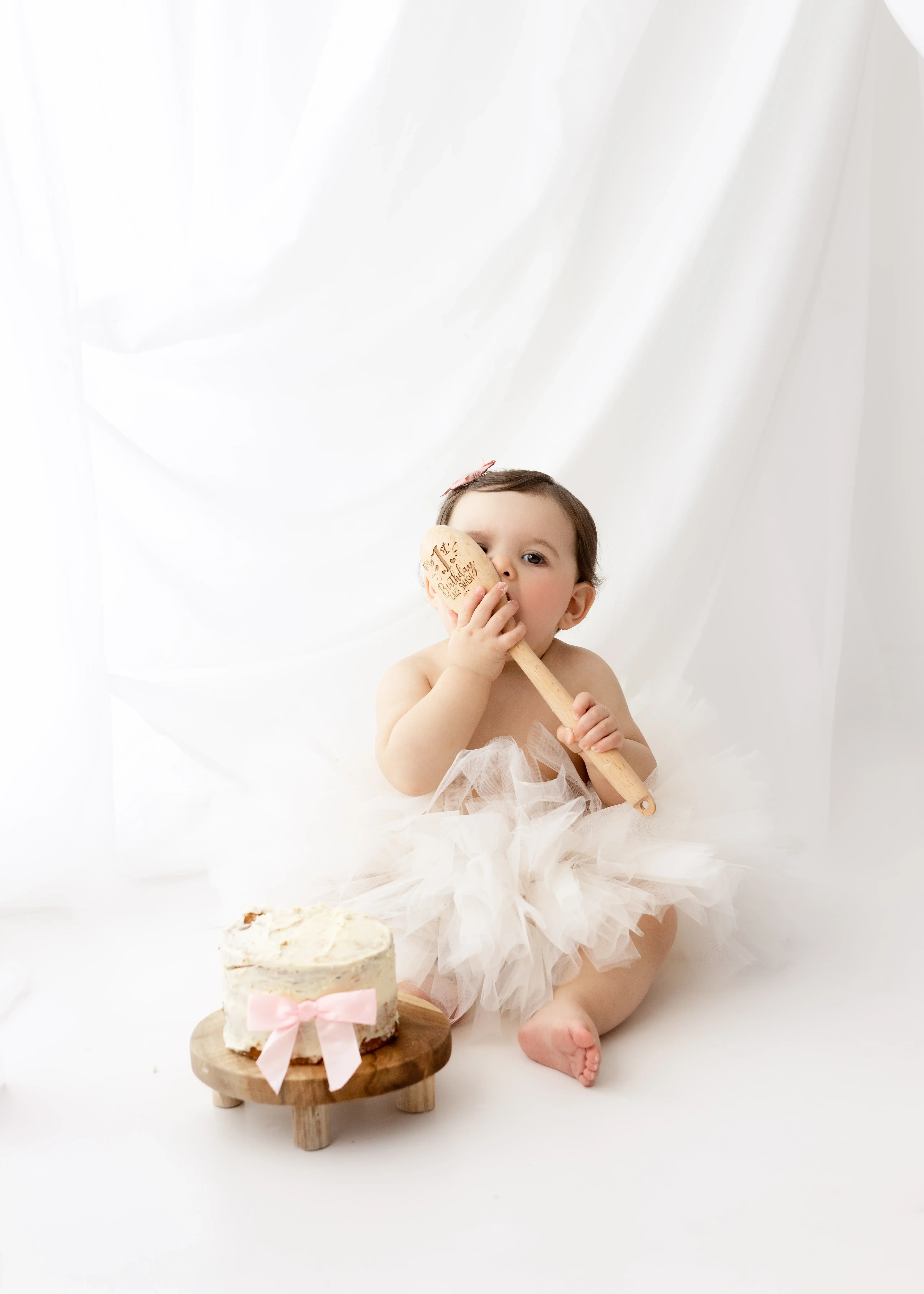 Little girl wearing a blush-pink romper, sitting next to a large pink number “1” balloon in a bright studio, capturing a simple and sweet first birthday milestone moment.