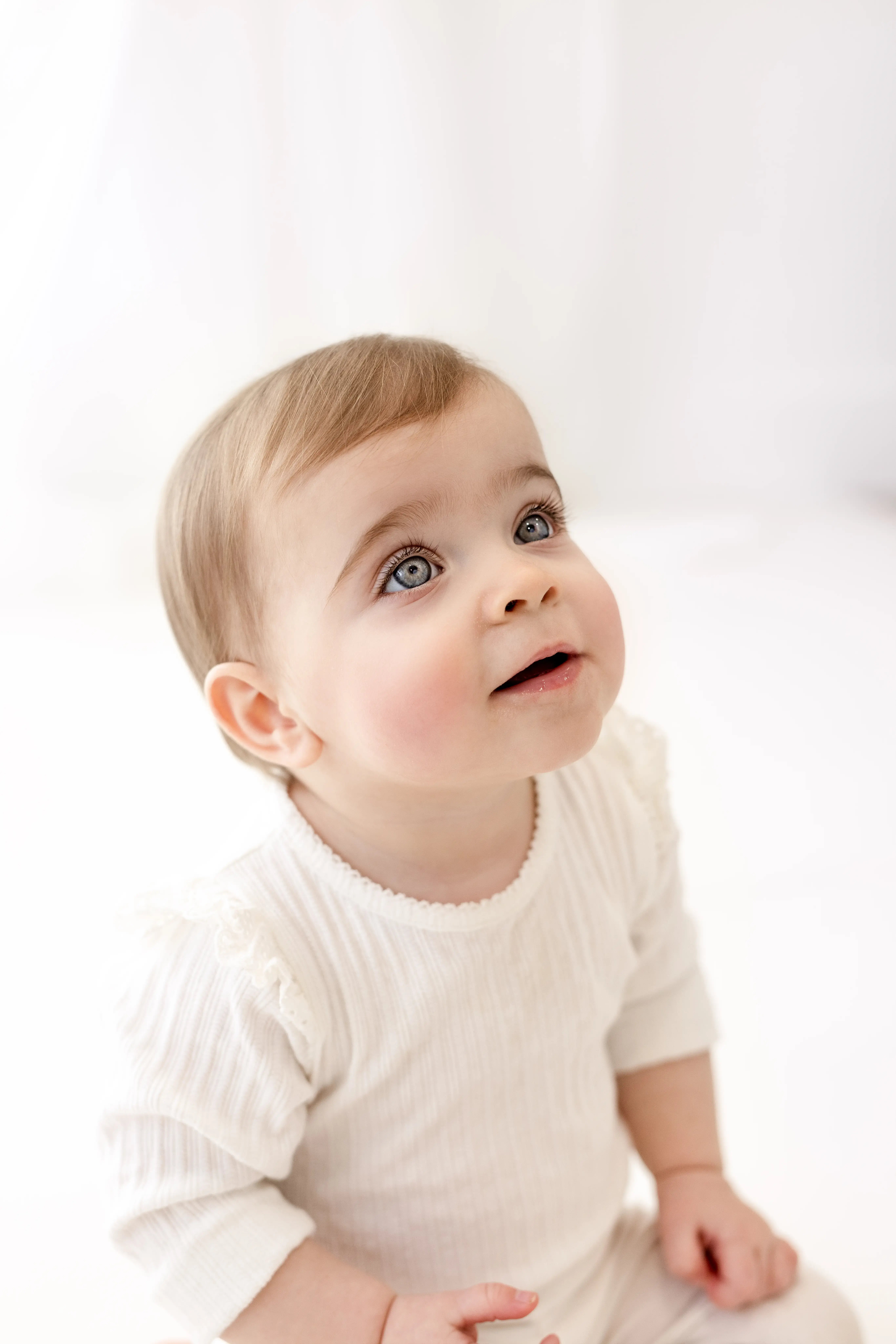 Close-up portrait of a bright-eyed baby girl in a cream-coloured outfit, gazing upward in a softly lit studio, capturing a sweet first birthday milestone moment.