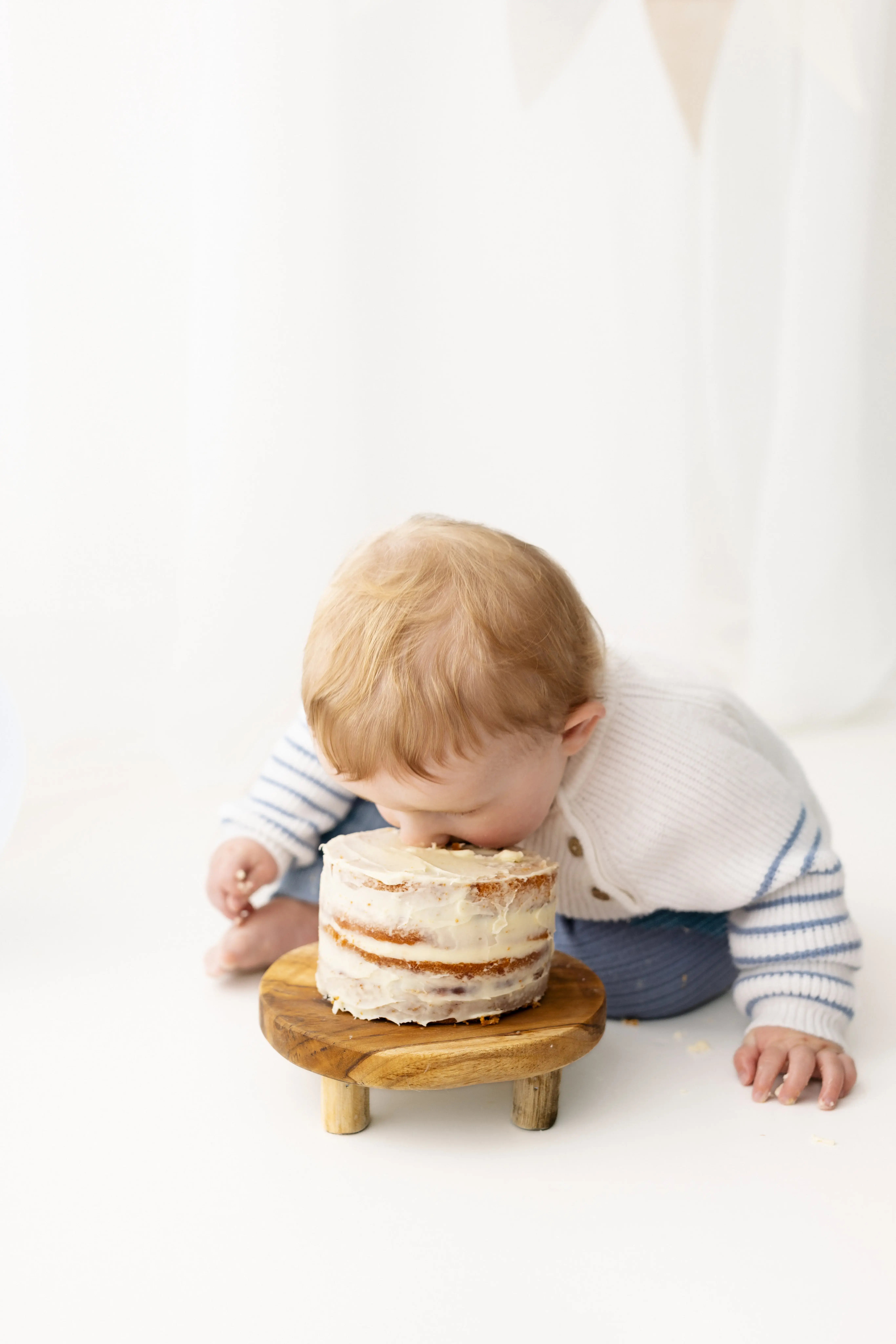 Giggly little boy in a striped knit jumper, leaning forward to take a bite directly from his frosted cake during an adorable first birthday cake smash.