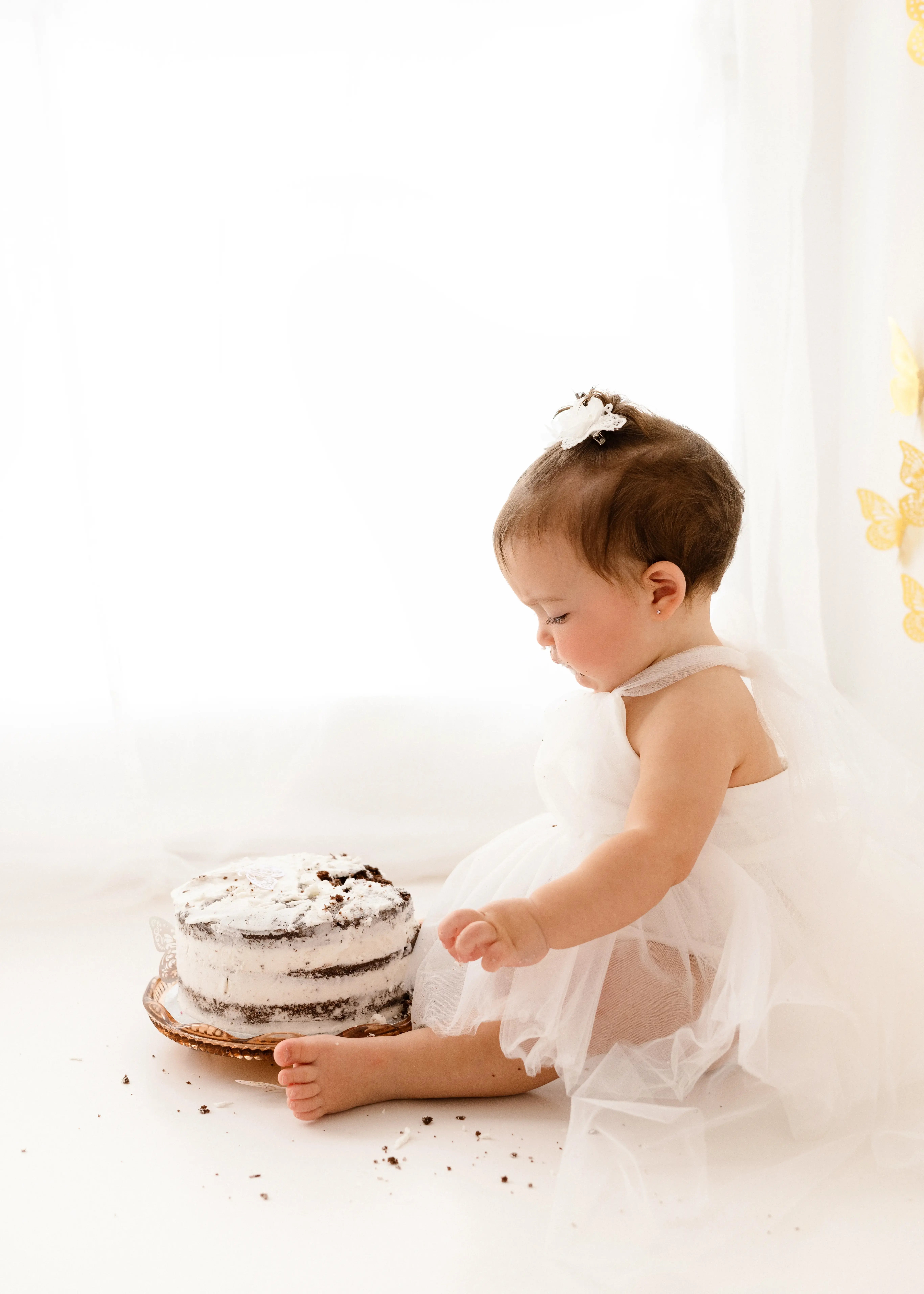 Baby girl in white tulle dress sitting on the floor beside a white buttercream cake during a cake smash first birthday photoshoot.