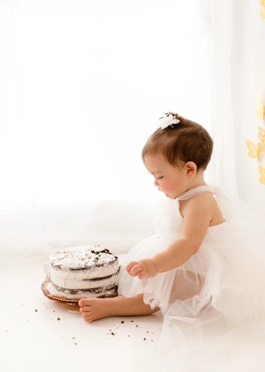 Baby girl in white tulle dress sitting on the floor beside a white buttercream cake during a cake smash first birthday photoshoot.