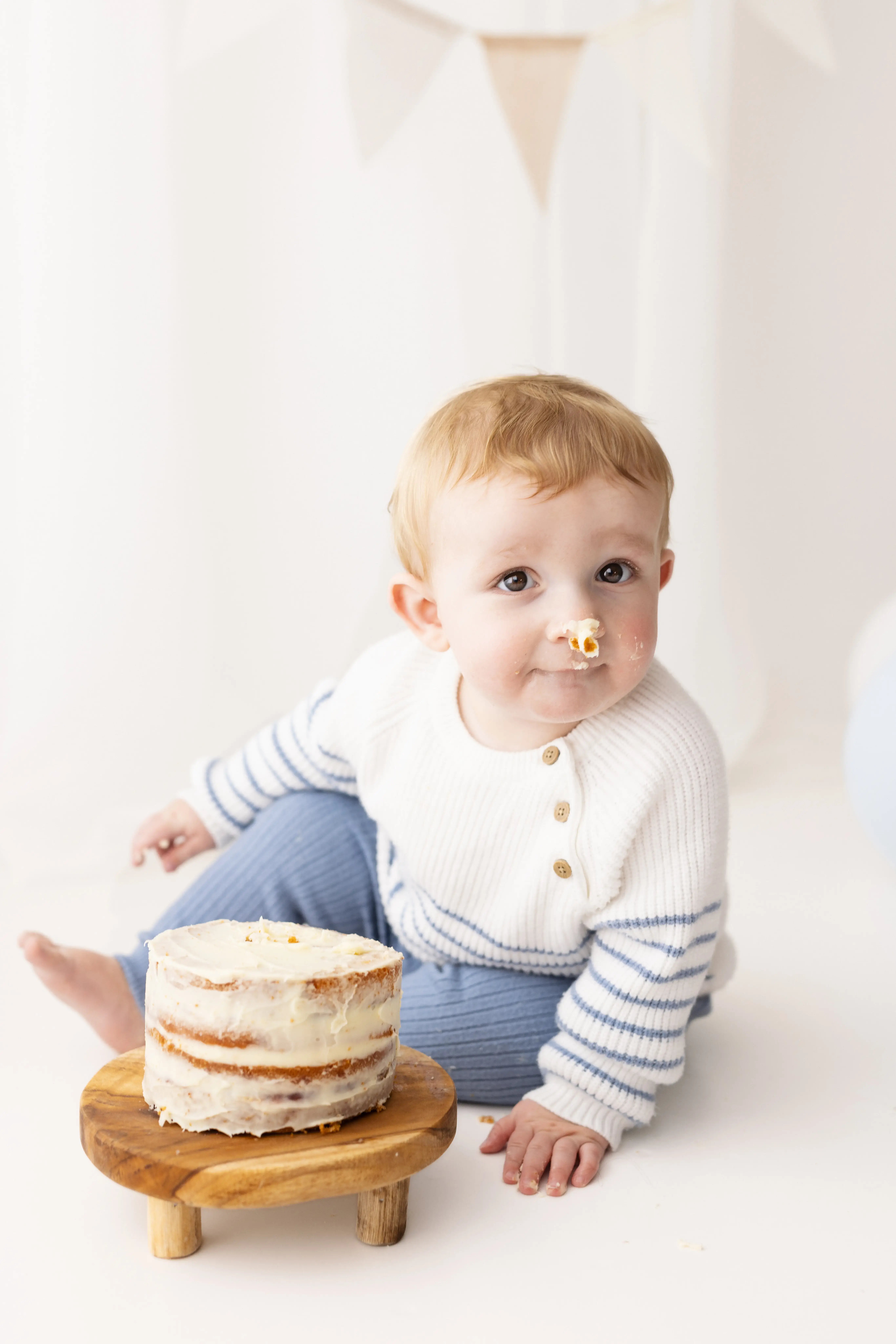 Toddler boy with cake frosting on his nose, seated beside a semi-naked frosted cake and wearing a cosy striped jumper, creating a fun, playful first birthday memory.