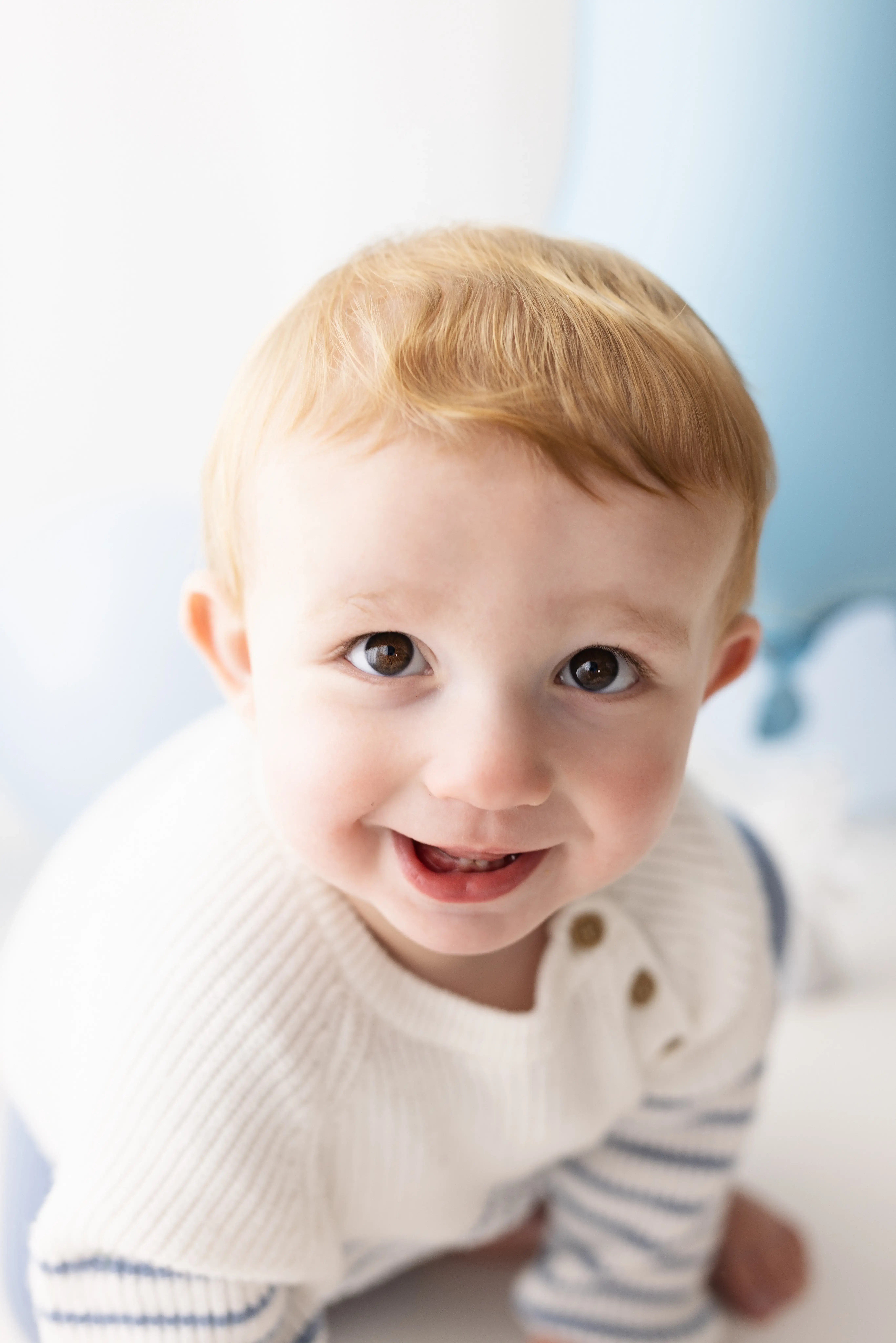 Cheerful toddler boy in a cosy white-and-blue sweater, glancing at the camera with a big grin, celebrating his first birthday in a bright, minimalist studio.