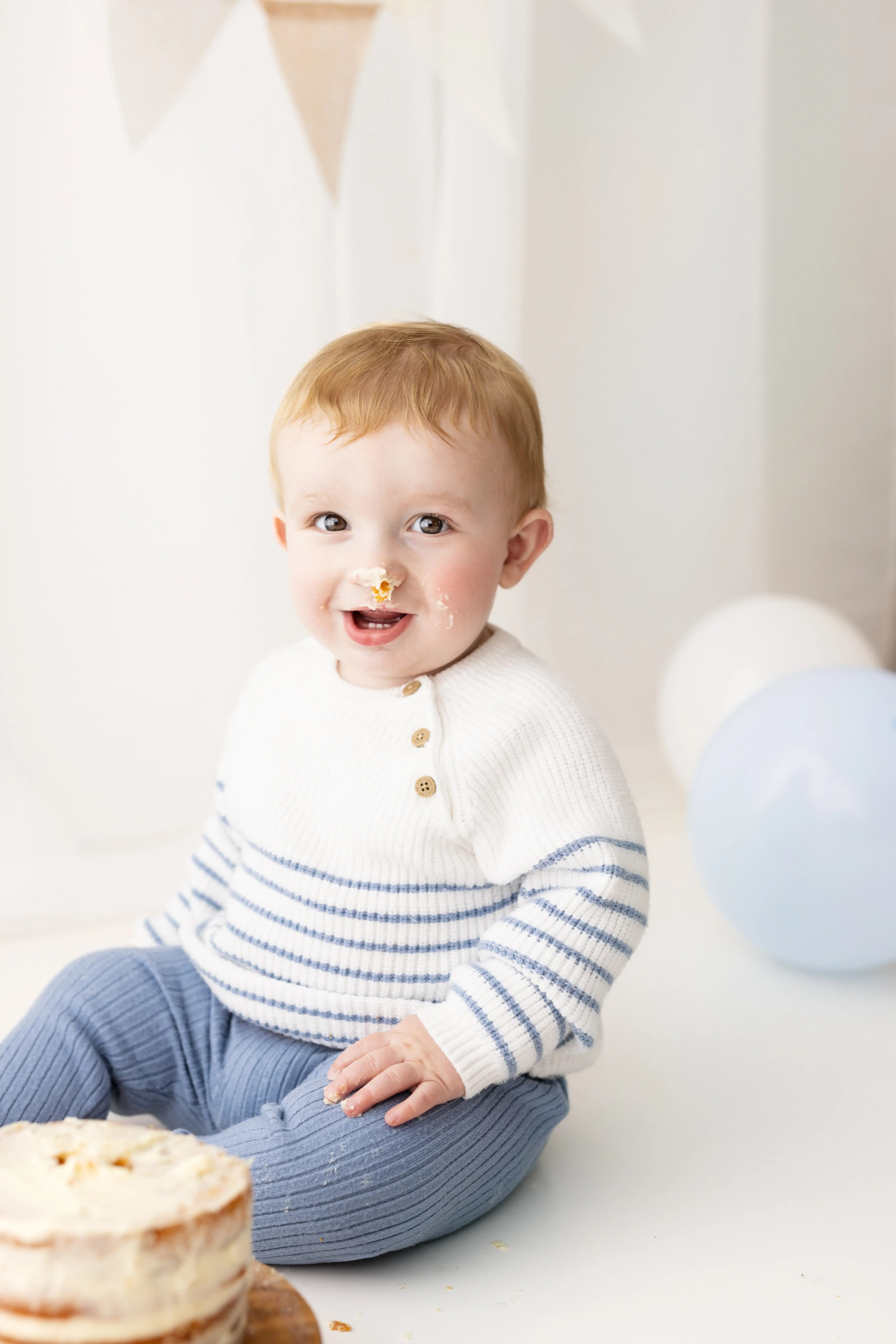 Cheerful toddler wearing a cosy white-and-blue striped sweater, sporting cake frosting on his nose as he enjoys his first birthday cake smash in a light-filled studio.
