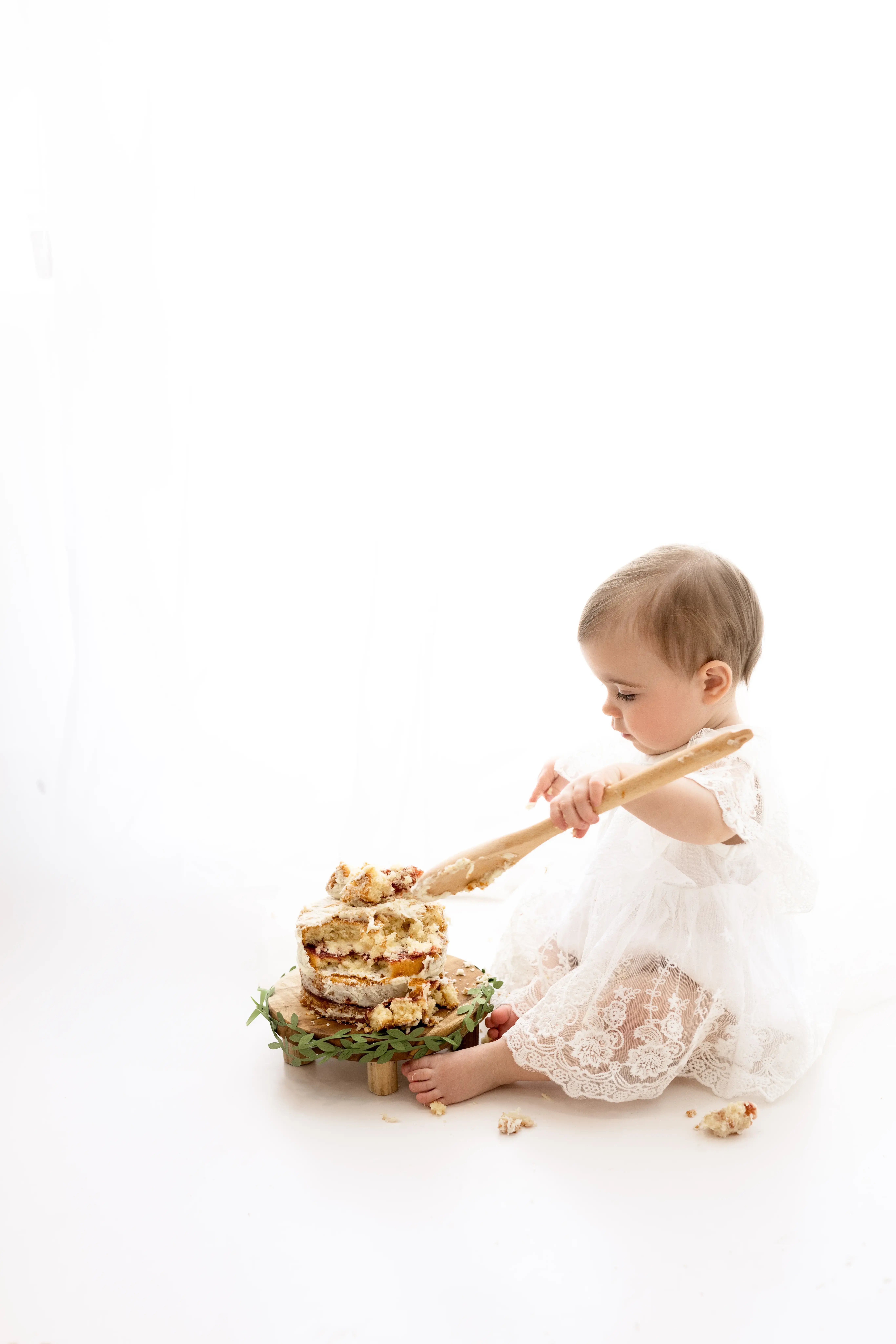 Baby girl in a delicate white lace dress enthusiastically smashing a rustic layered cake with a wooden spoon, celebrating her first birthday in a soft, all-white studio setting.