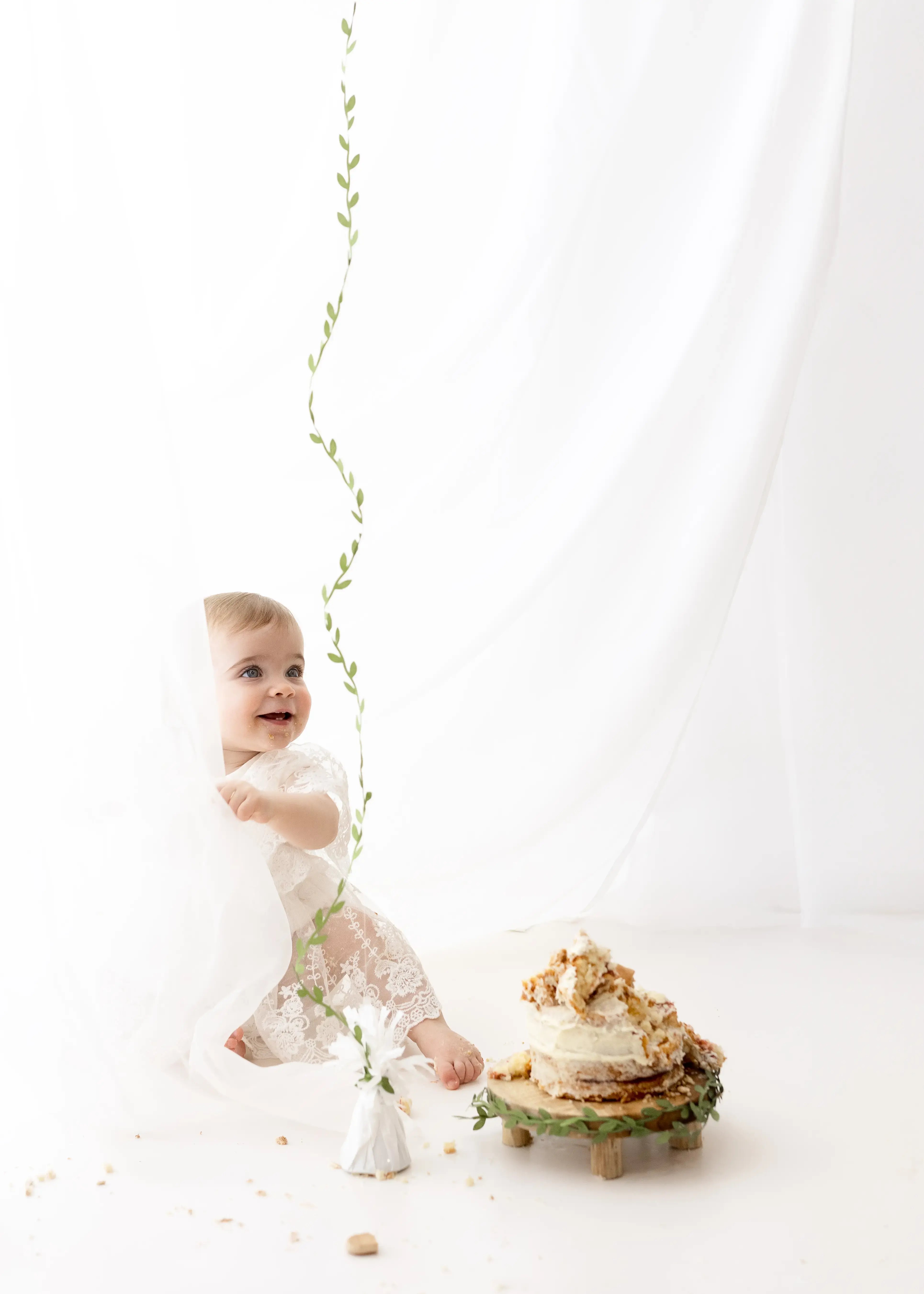 Smiling infant dressed in a lace romper, playfully peeking from behind a sheer curtain while admiring a large white balloon and her half-eaten cake during a cake smash session.