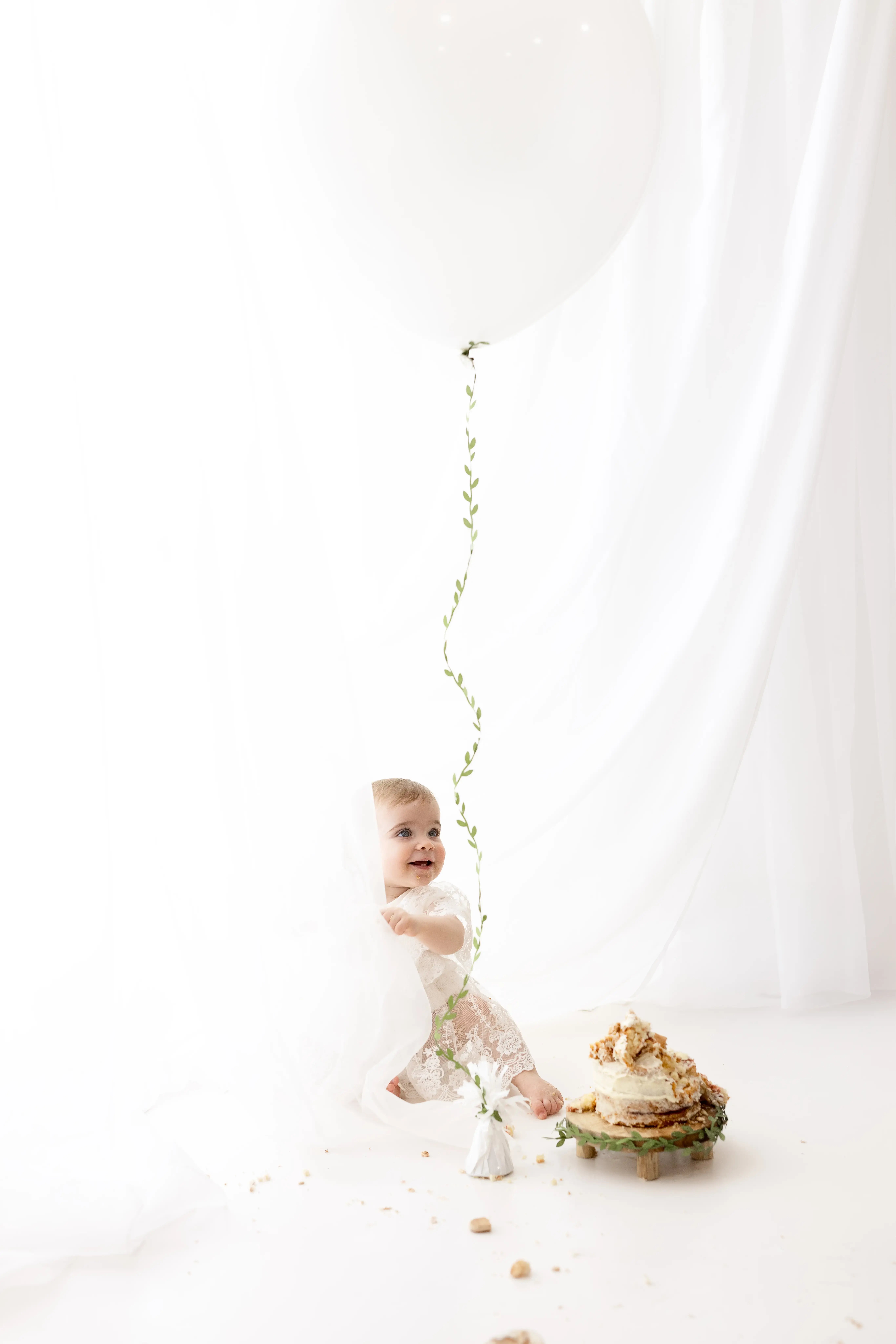 Smiling one-year-old in a pretty white lace romper, peeking out from behind a sheer curtain and playing with a large white balloon while her cake sits half-smashed on a wooden stand.