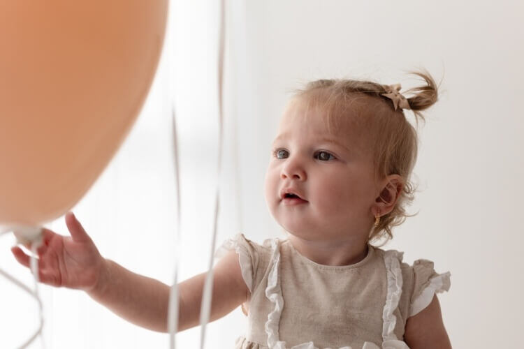 Photograph showing a young girl at a cake smash photography shoot, pictured playing with her balloons wearing a dress