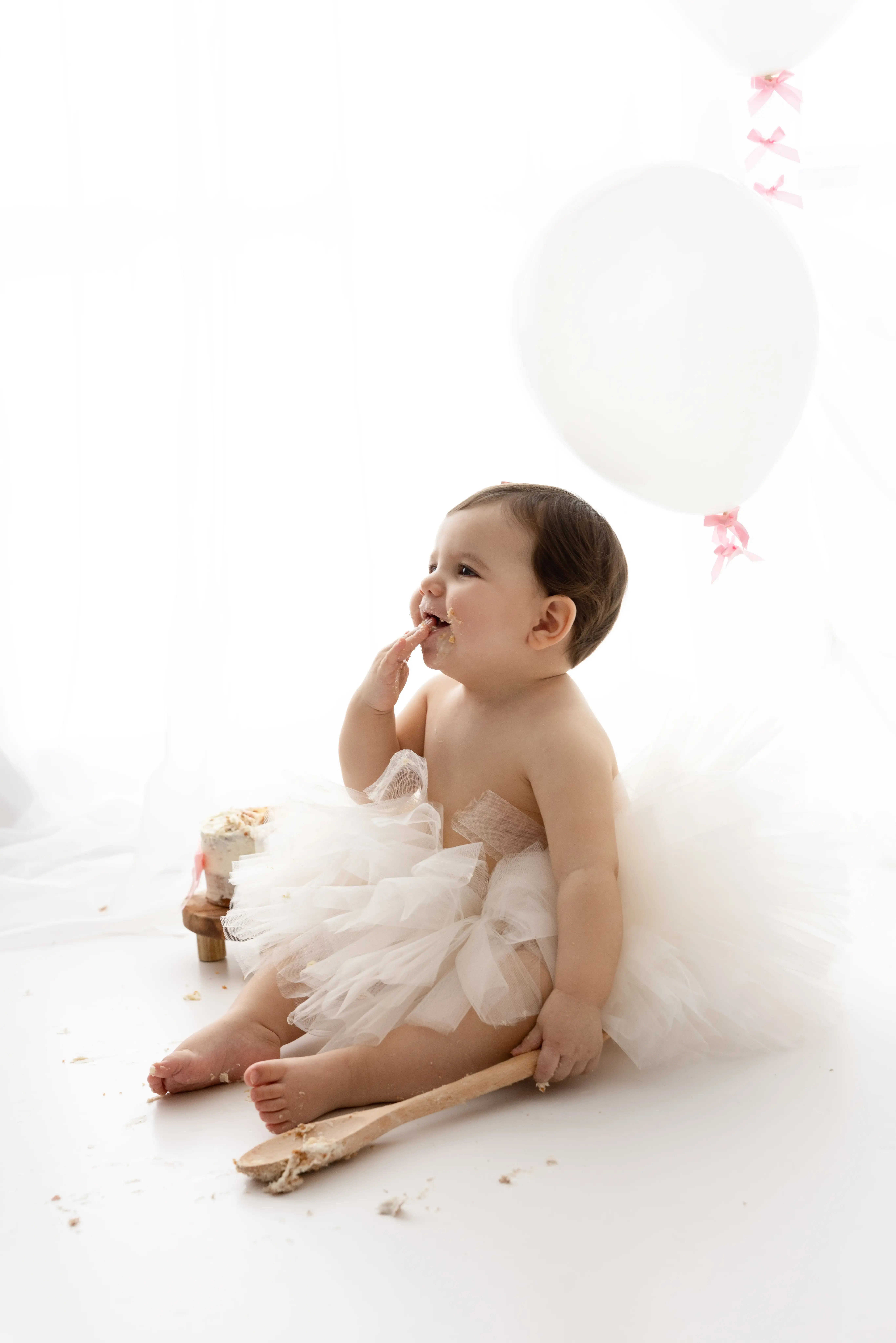 Smiling baby in a white tulle skirt, happily tasting her first birthday cake with a wooden spoon, surrounded by soft white balloons in a naturally lit photography studio.