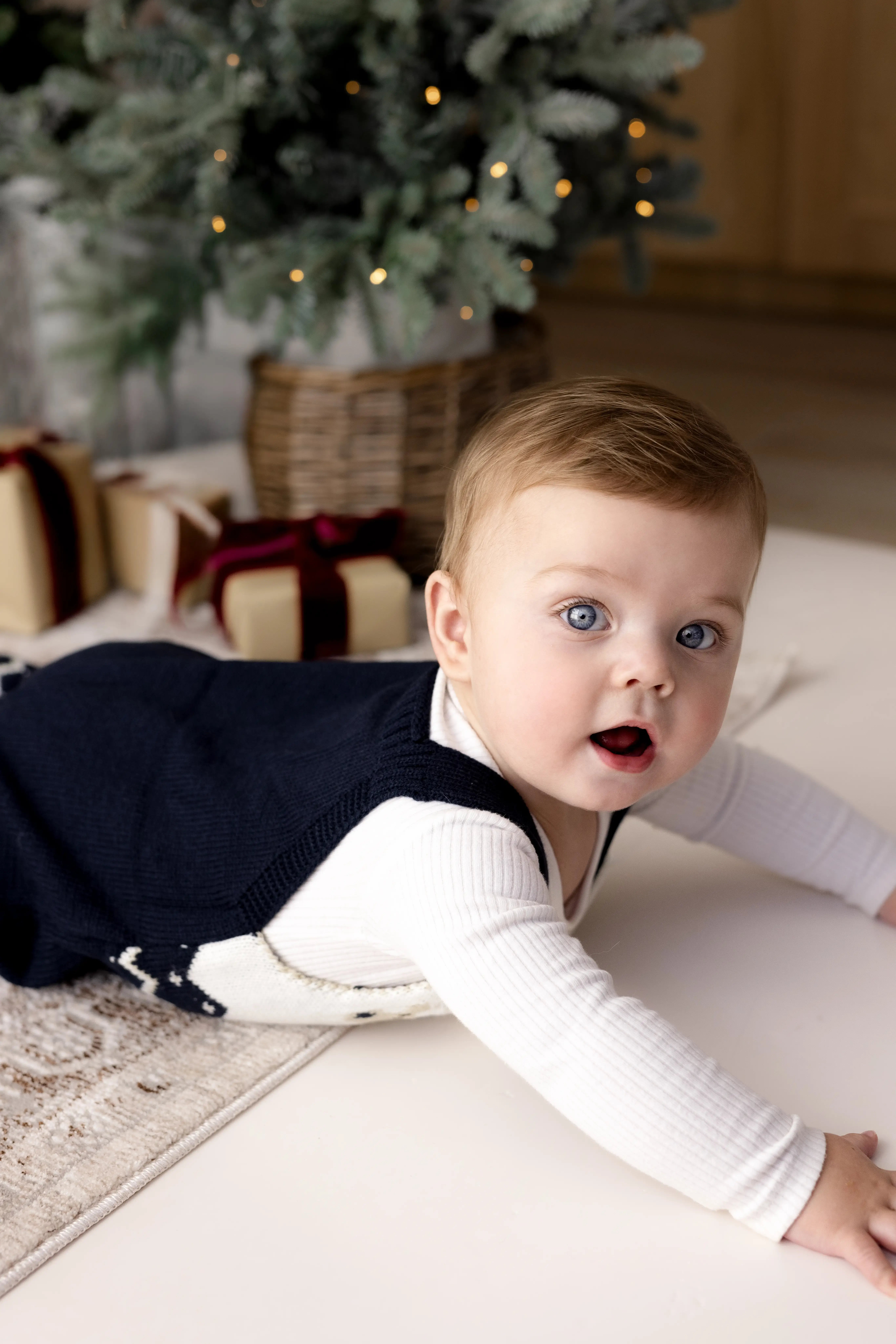 Curious baby crawling on a white studio floor, wearing a navy reindeer jumper with gift boxes and frosted Christmas trees in the background for a cosy holiday photo.