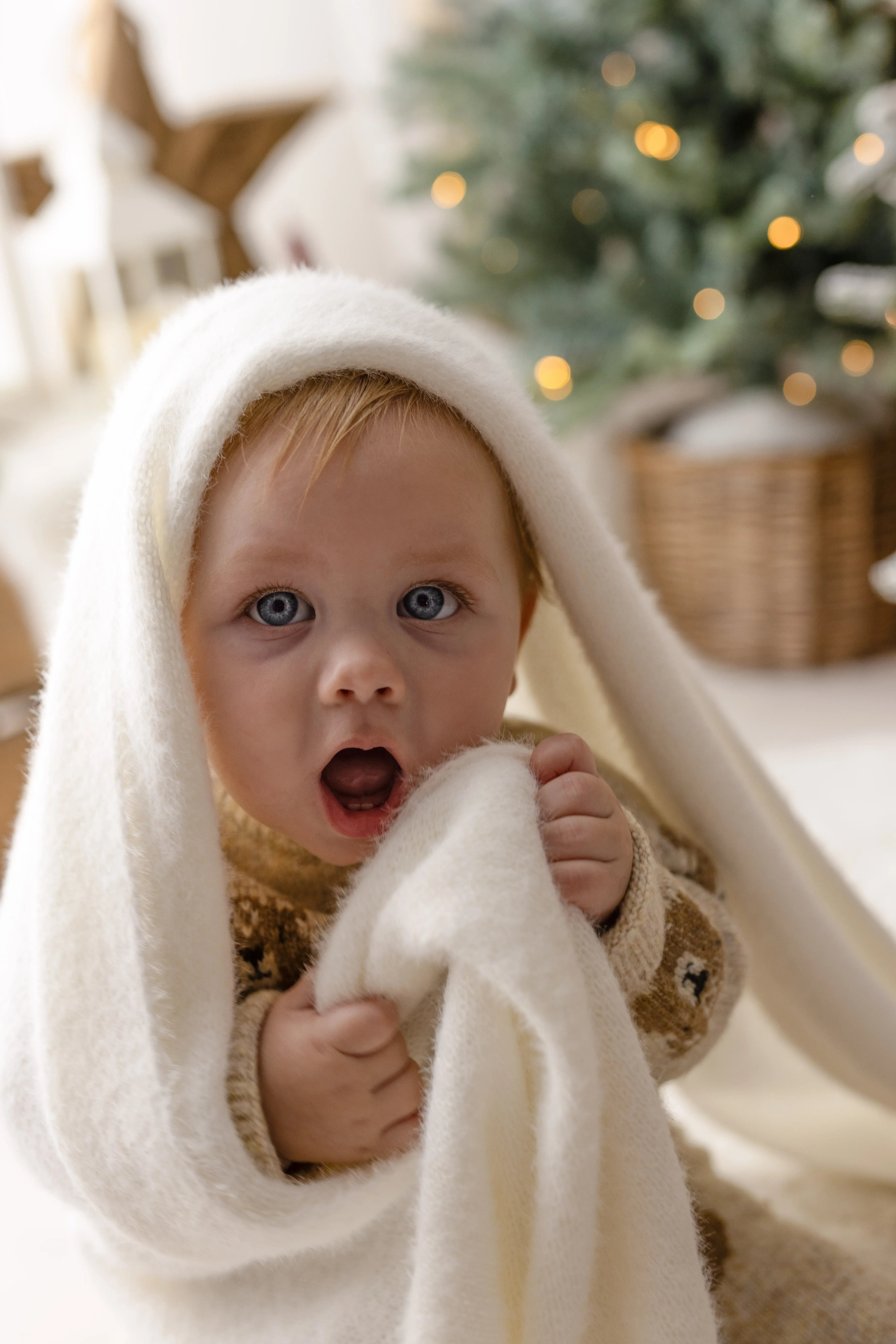 Wide-eyed baby boy peeking out from under a fluffy white blanket, surrounded by warm fairy lights and Christmas décor for a magical holiday mini session.