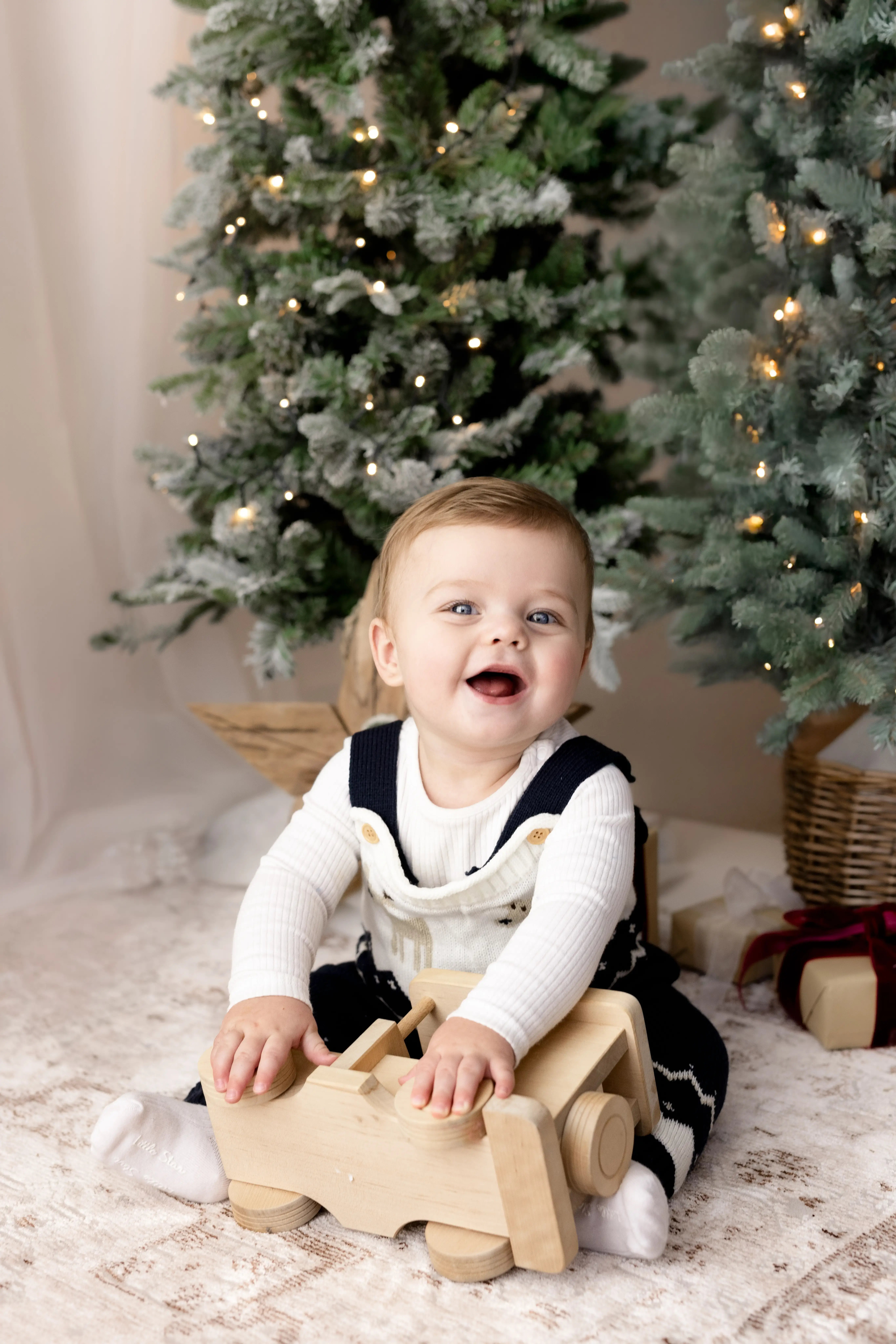 Giggling baby boy in a winter-themed knit outfit, happily playing with a wooden toy truck among snowy Christmas trees and presents, creating a warm festive feel.