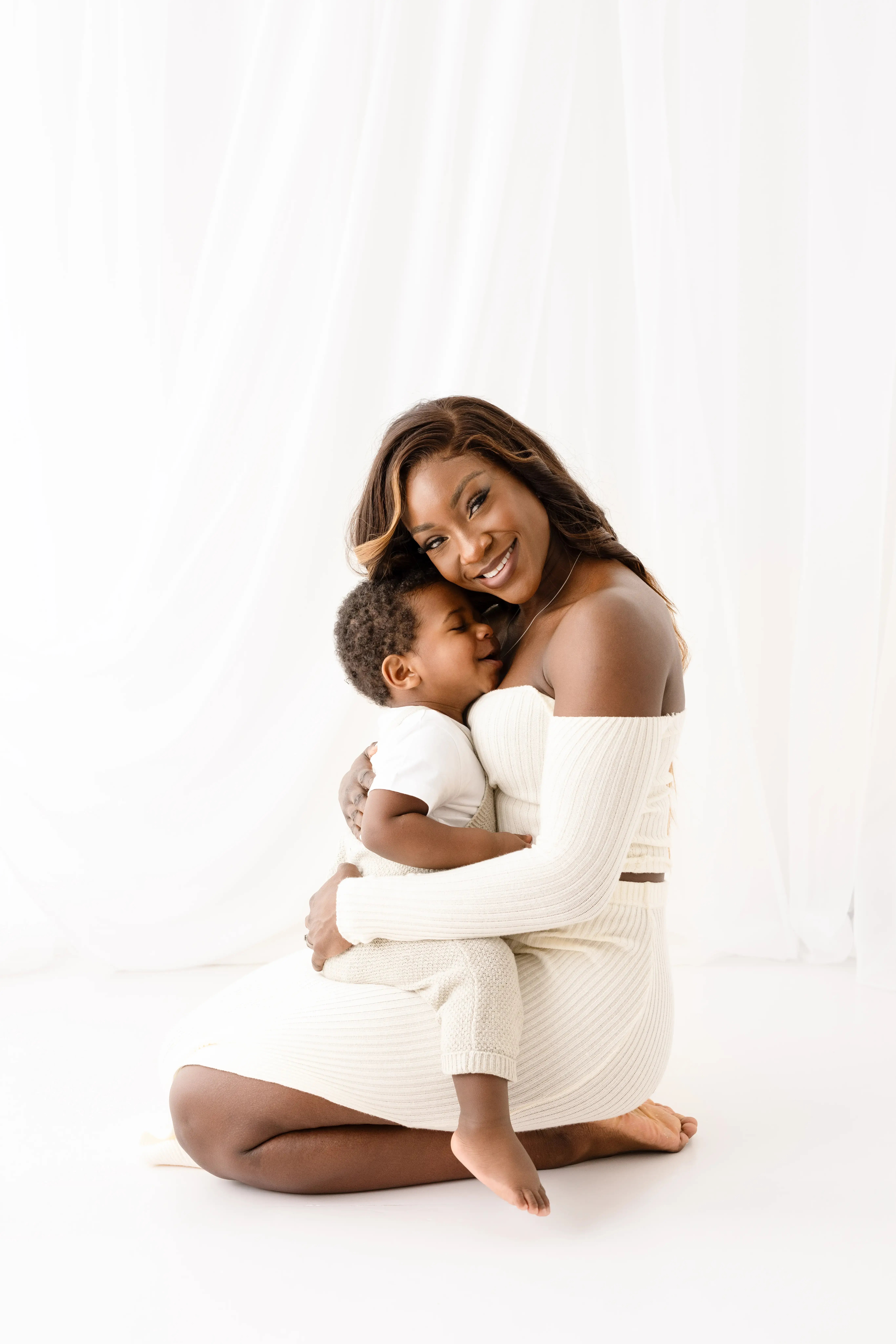Loving mother in a cream off-the-shoulder knit dress, kneeling and cuddling her young son as he rests his head on her chest, both dressed in cosy neutral tones against a white backdrop.