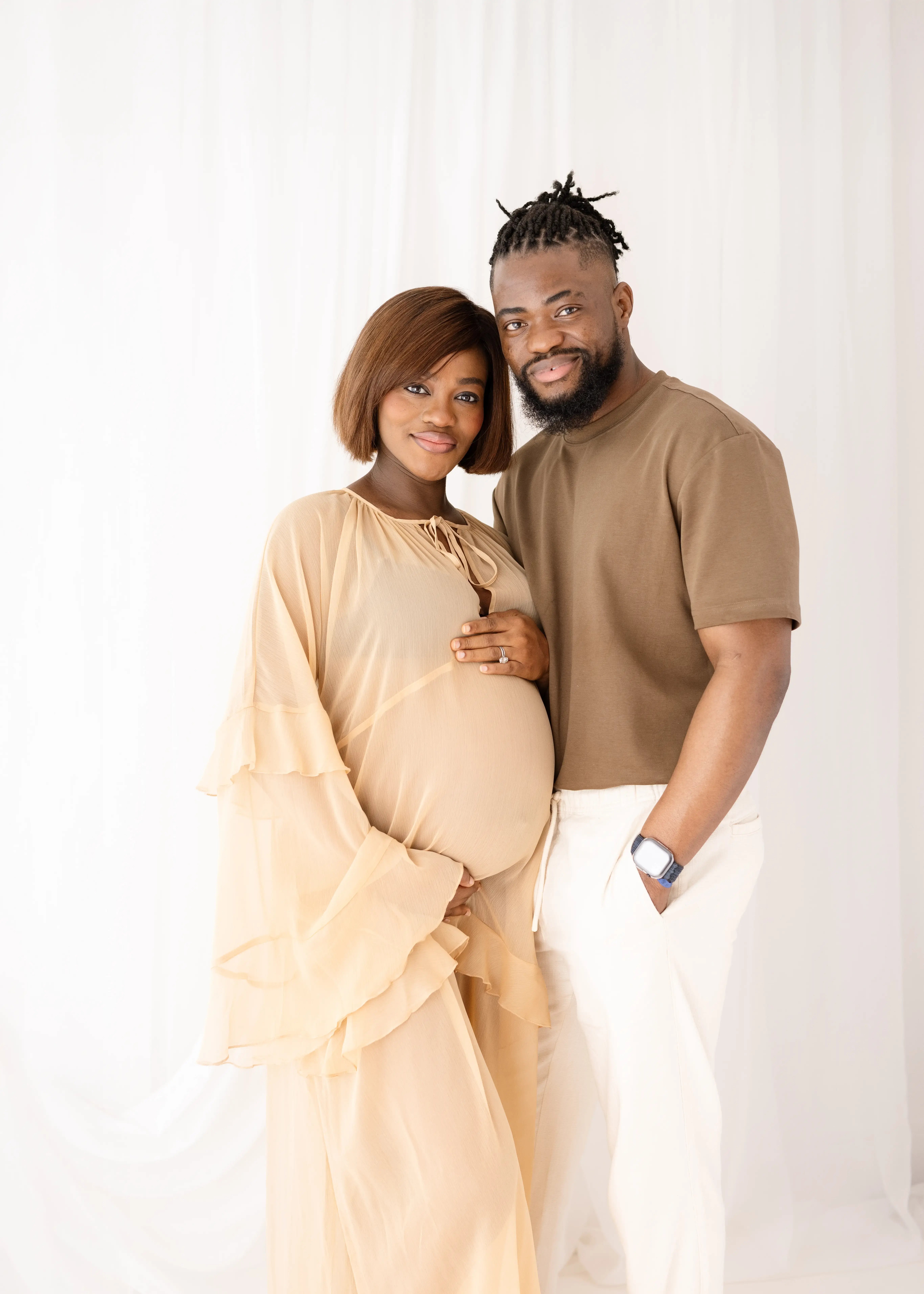 Expectant couple smiling warmly, with the pregnant woman in a flowing beige gown and the man in a brown t-shirt and cream trousers, standing close together in a white studio setting.