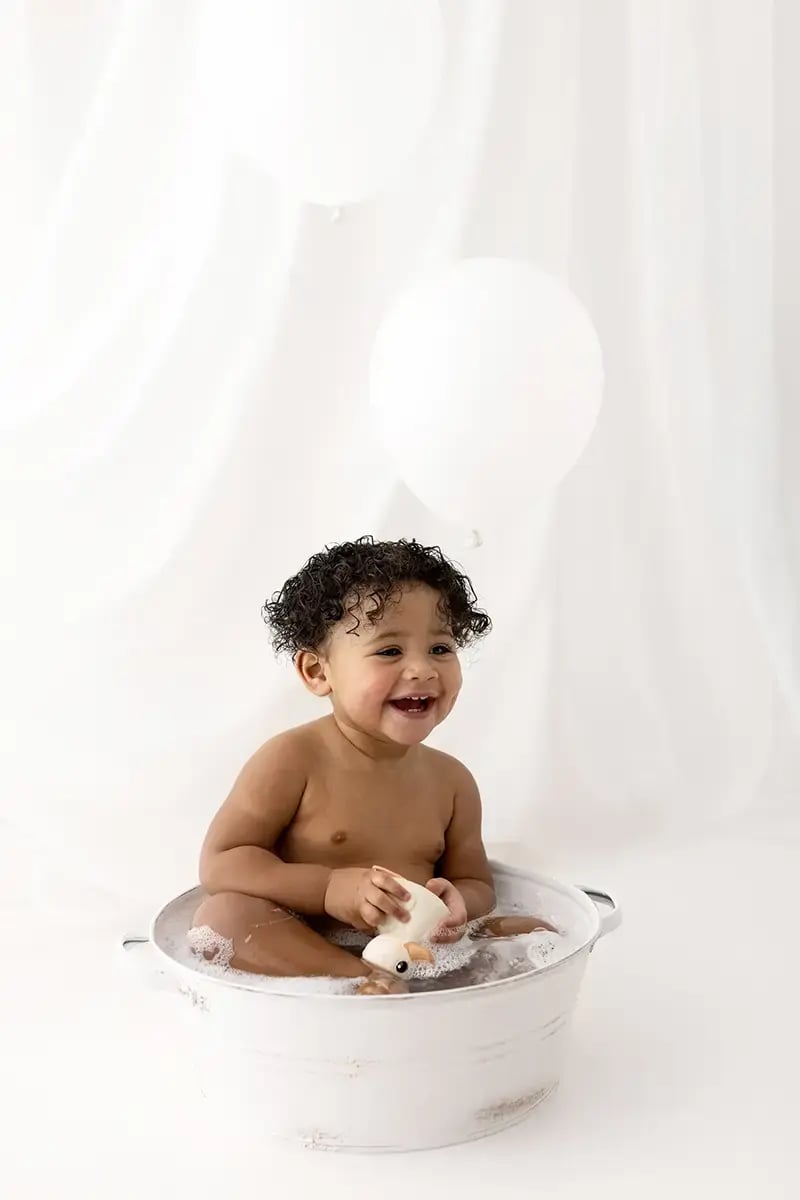Smiling baby with curly hair enjoying a bubble bath in a white metal tub, holding a white rubber duck, set against a soft white background with white balloons.