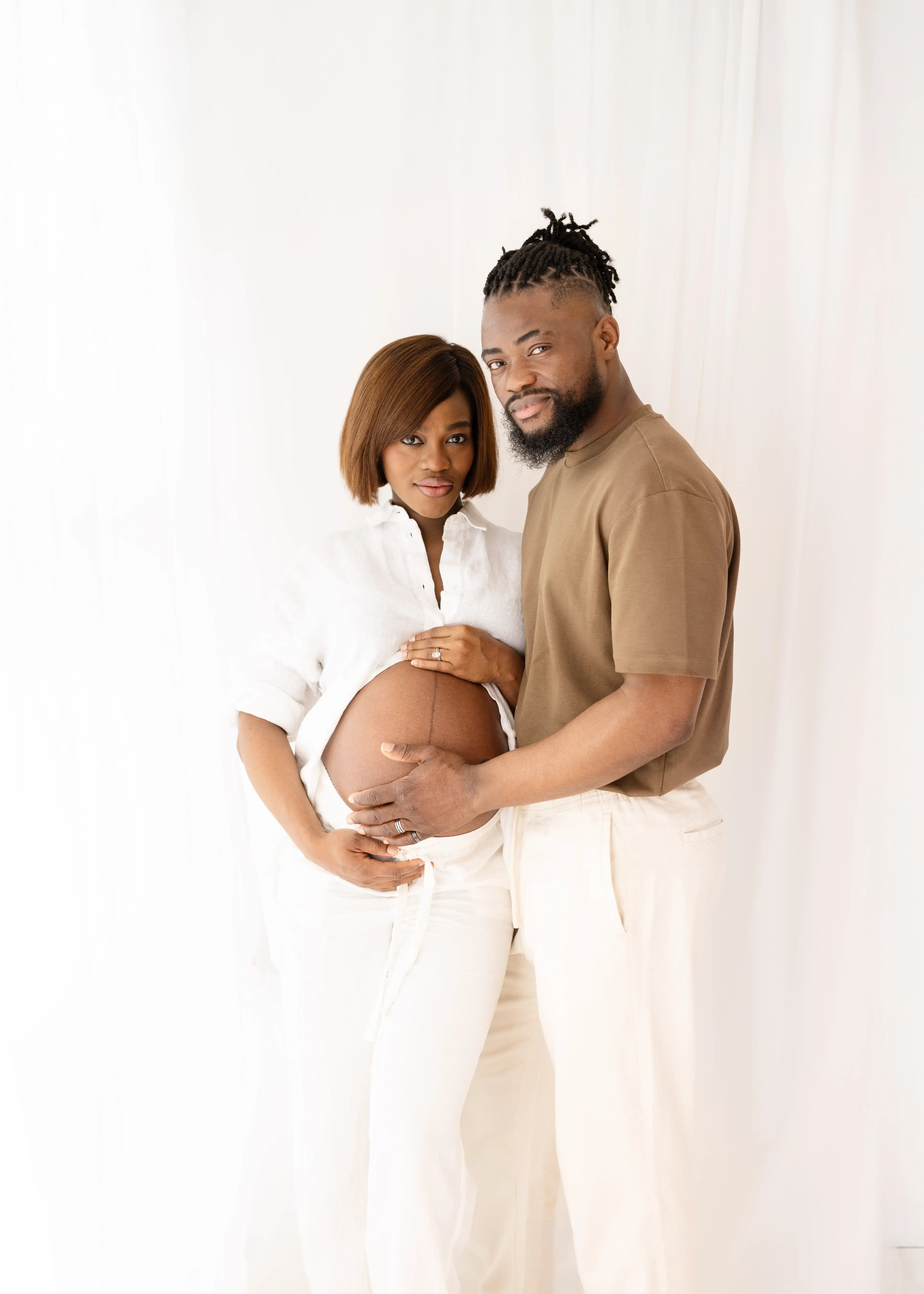 Expectant couple embracing closely, both placing their hands on the woman's exposed bump as she wears white and he wears a brown top with cream trousers, set against a minimalist white background.