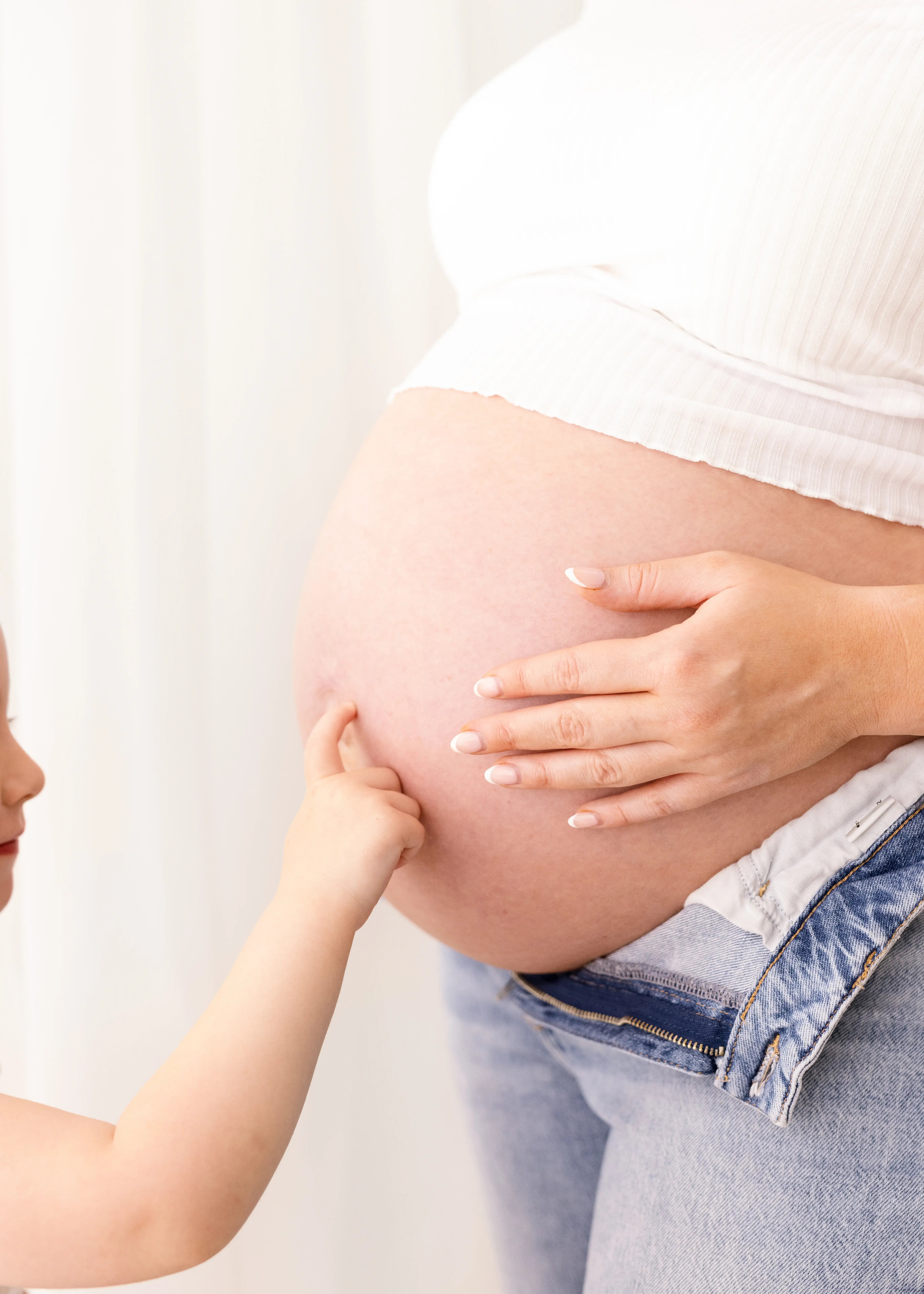Close-up of a child’s finger touching a pregnant belly while the mother’s hand rests protectively nearby, both wearing soft white and denim.