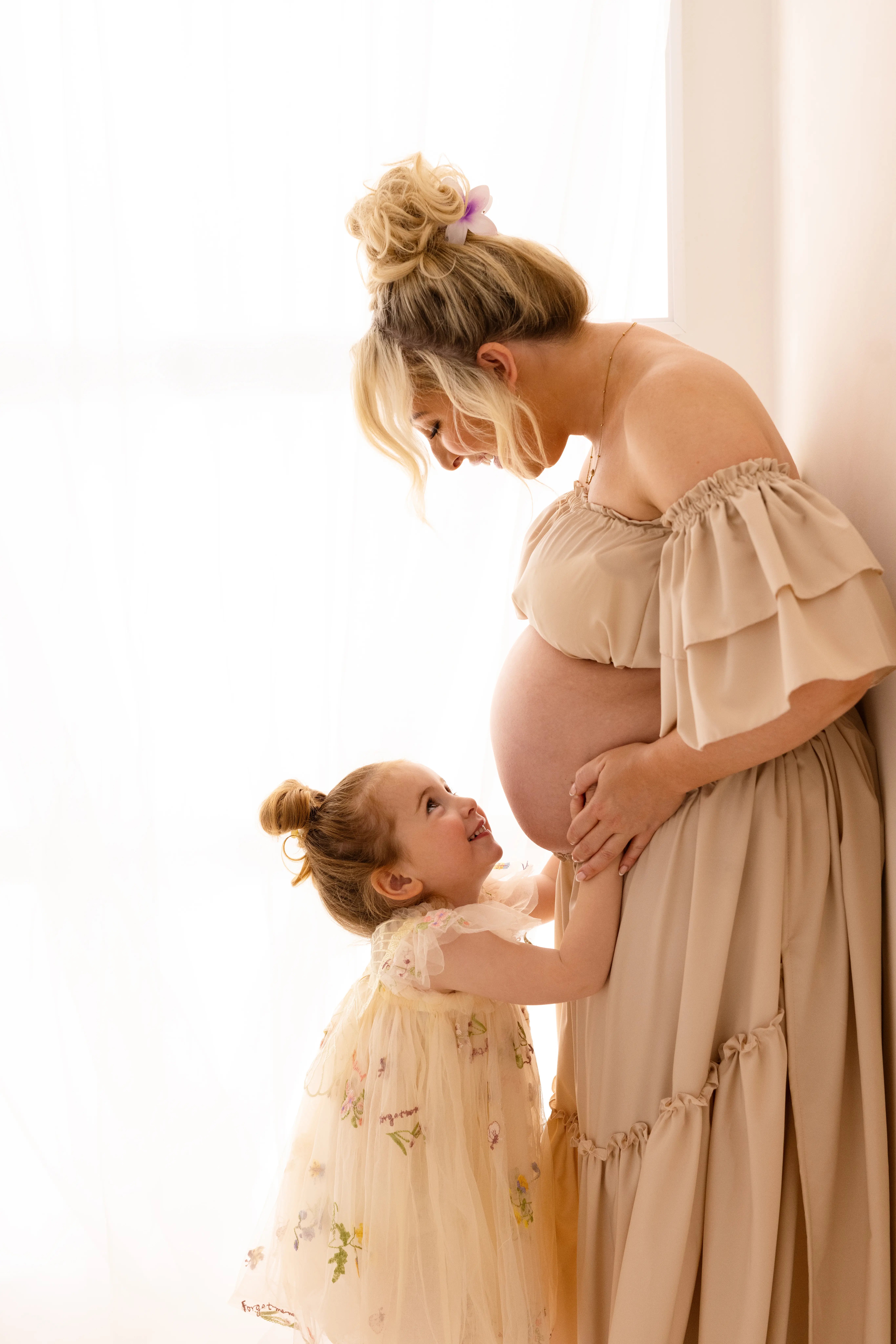 A young girl in a floral dress hugs her mum’s baby bump and looks up smiling, while her mother holds her hands in a softly lit studio.