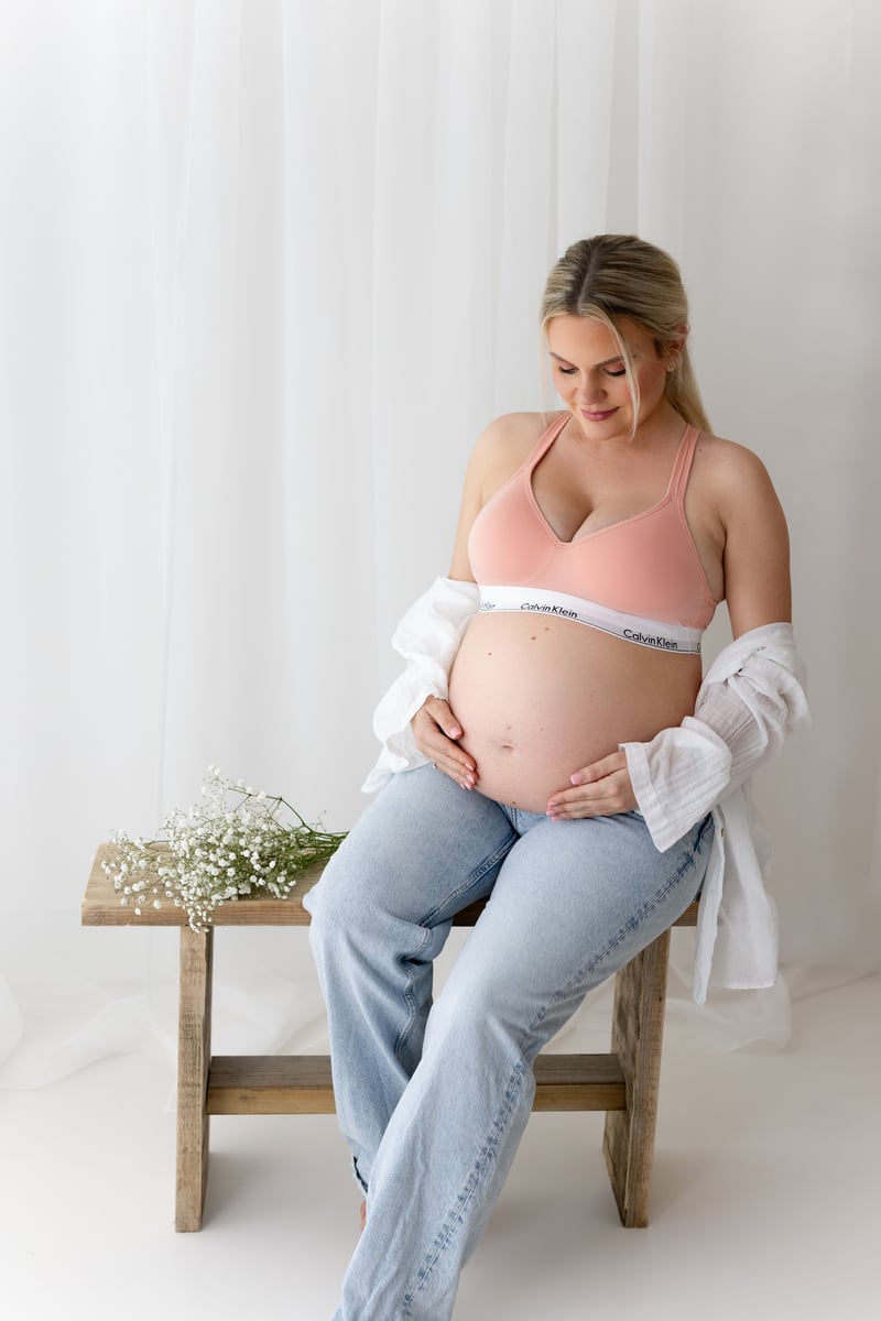 Photograph of a mum-to-be, pictured wearing denim jeans and sitting on a bench, looking at their bump