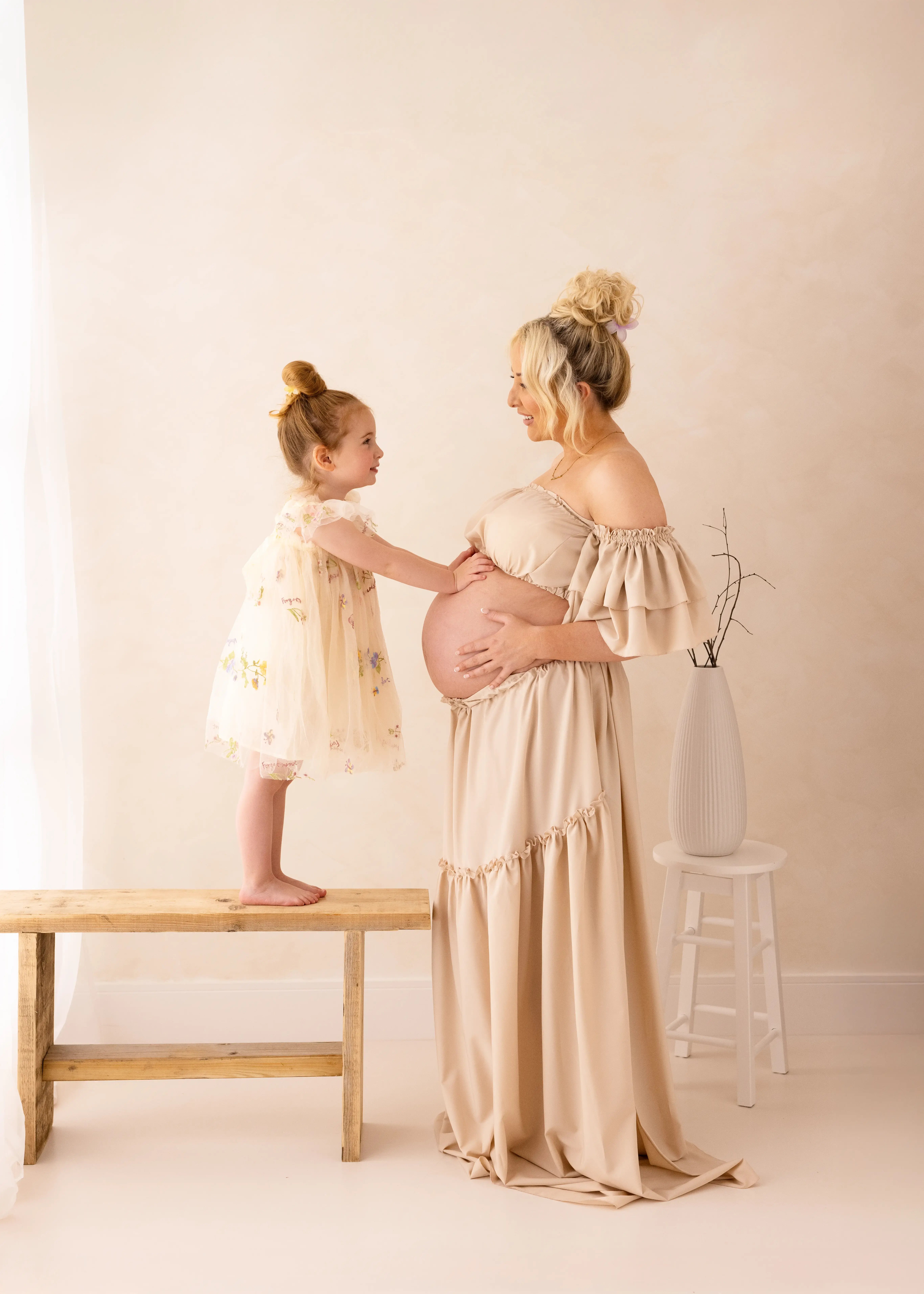 A pregnant woman in a flowing beige gown smiles at her young daughter who gently places her hands on her baby bump while standing on a bench.