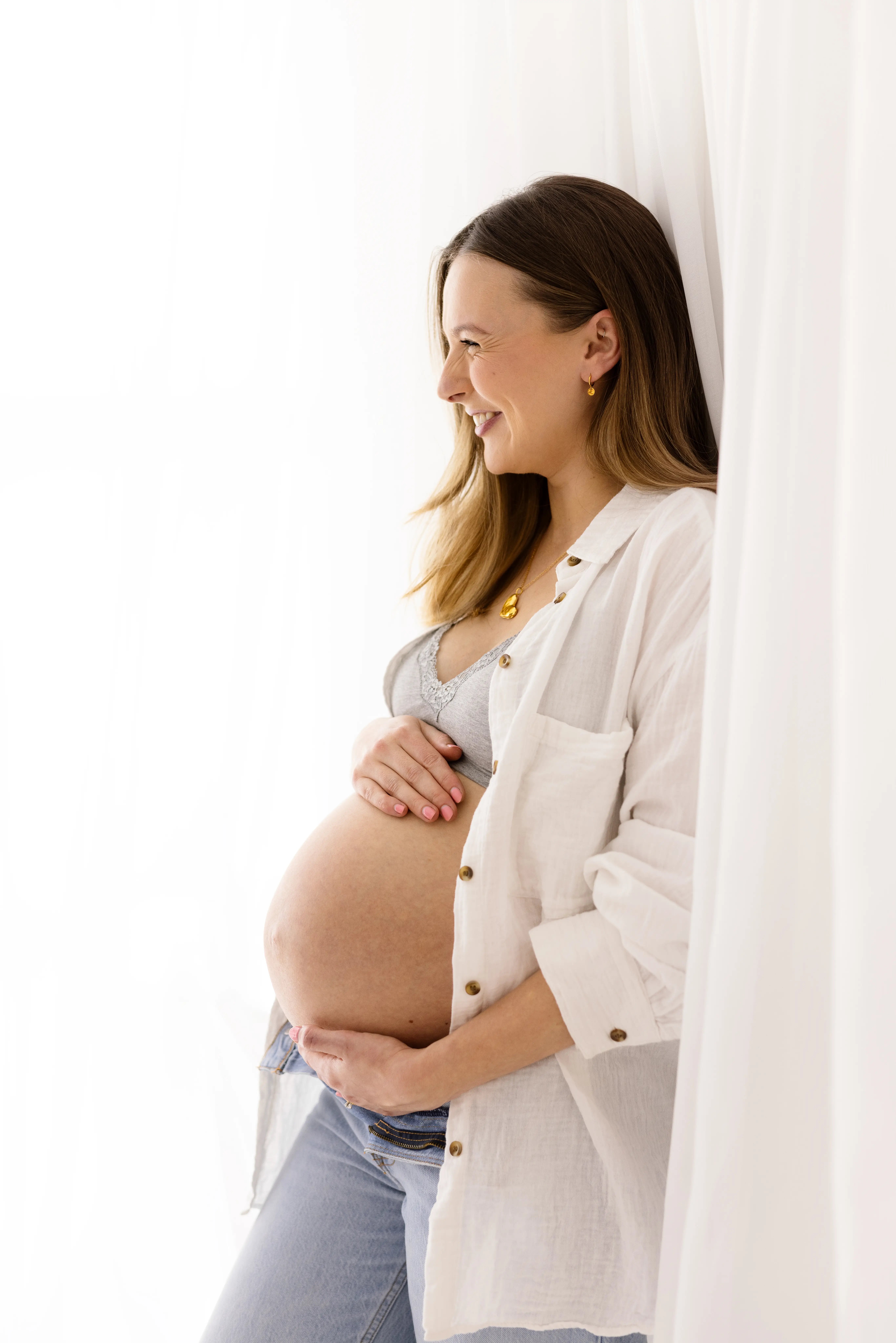Pregnant woman smiling as she holds her bump, wearing a white shirt and jeans beside sheer curtains in a maternity photoshoot.