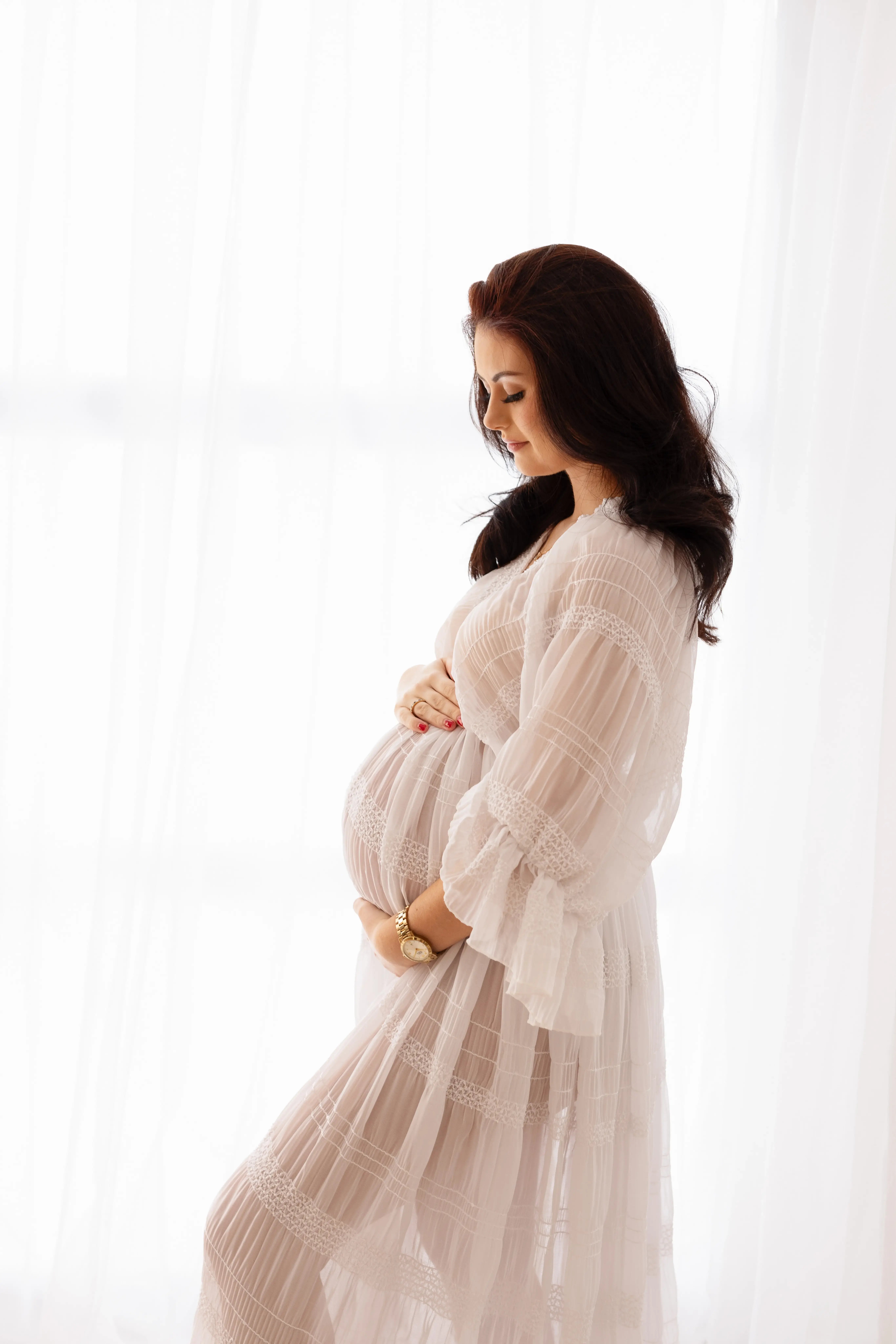 Graceful expectant mother in a sheer white lace gown, standing in a sunlit studio and tenderly holding her bump for an elegant, airy maternity photo session.