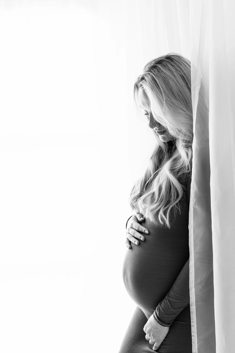 Black-and-white profile shot of a blonde mother-to-be in a form-fitting dress, softly touching her baby bump and standing behind a translucent curtain for a timeless maternity portrait.