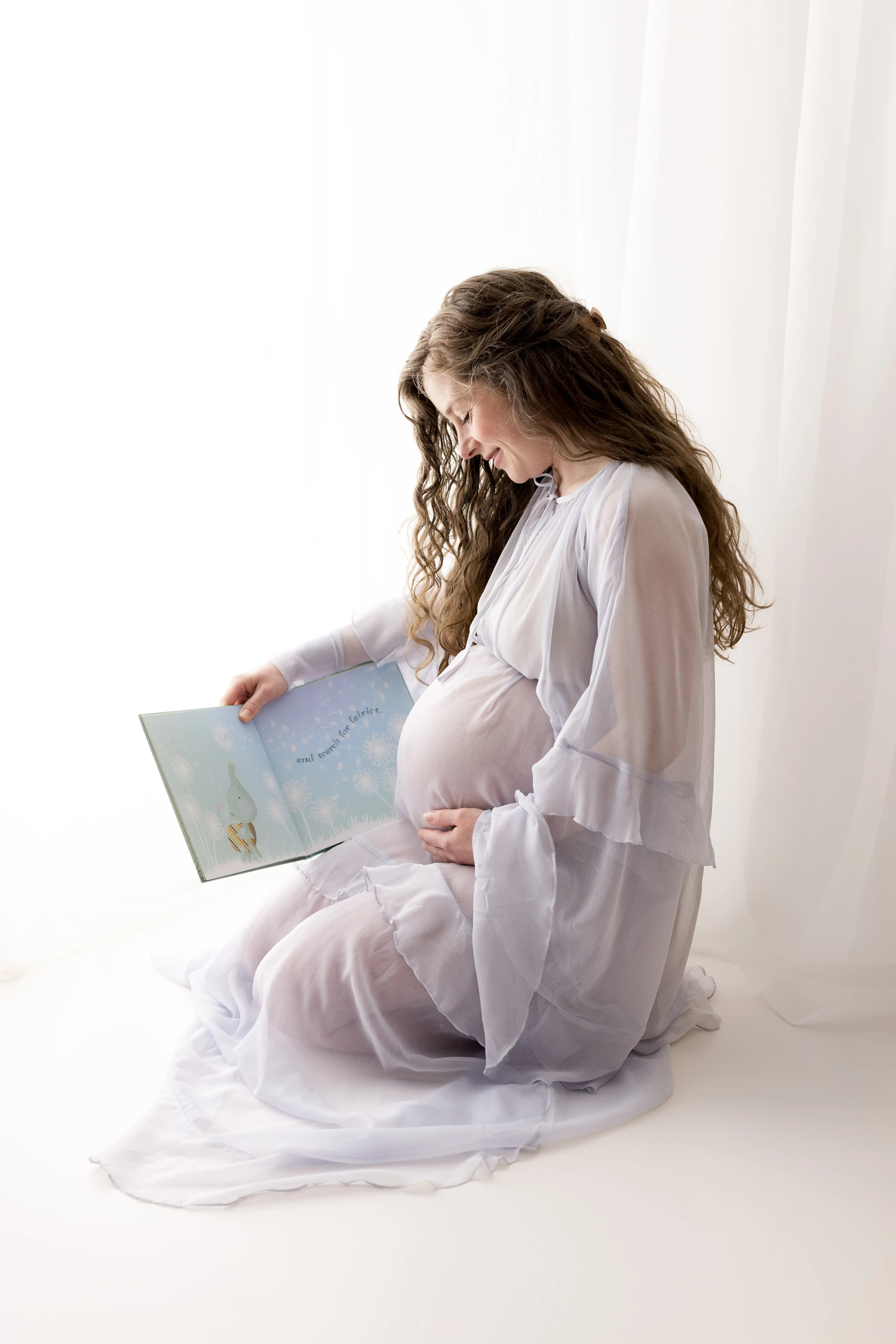 Seated pregnant woman in a sheer, flowing white robe reading a children’s storybook, highlighting a serene and gentle moment during a light and airy maternity photography session.