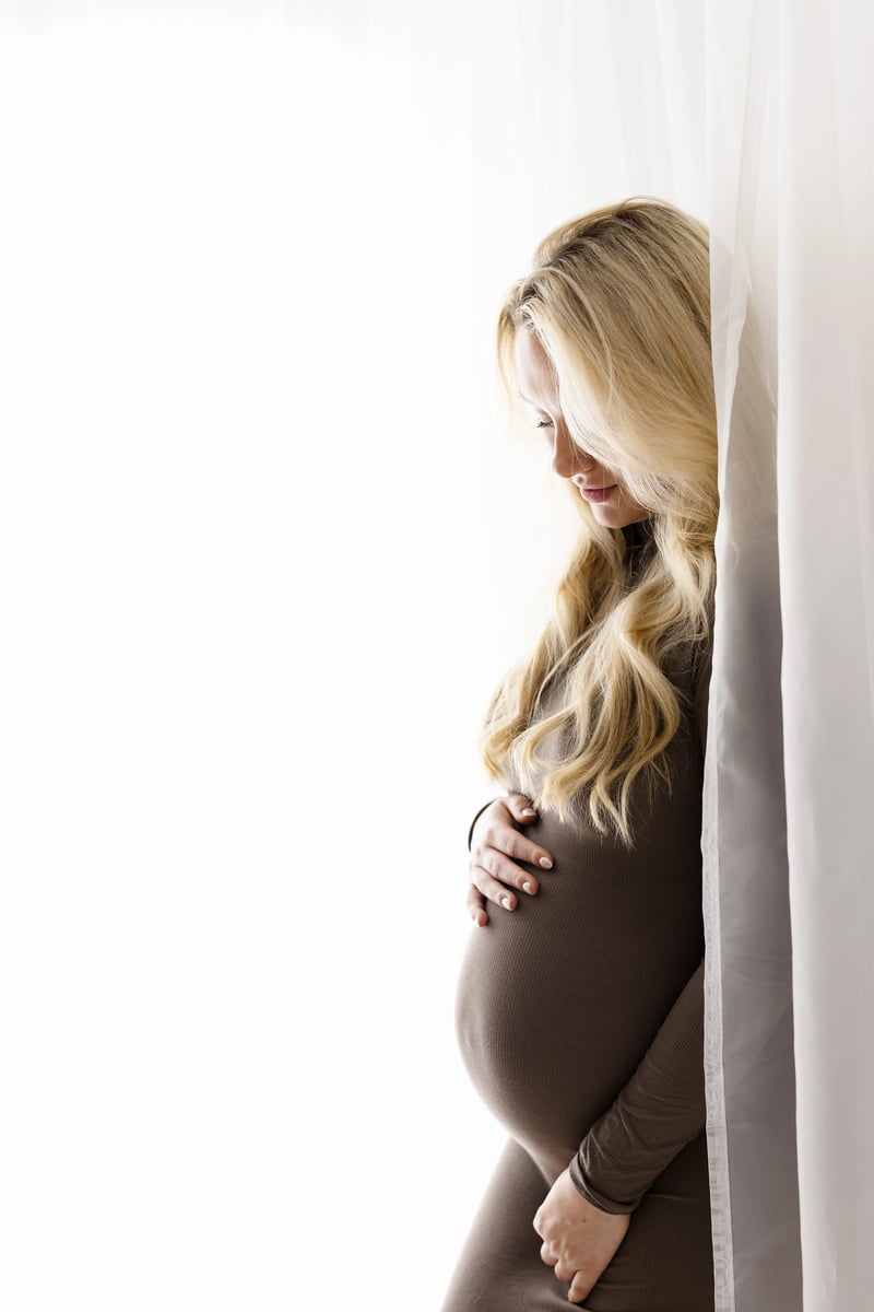 Side-profile of a pregnant blonde woman in a fitted brown maxi dress, gently cradling her baby bump while standing behind a sheer white curtain in a bright, minimalist studio.
