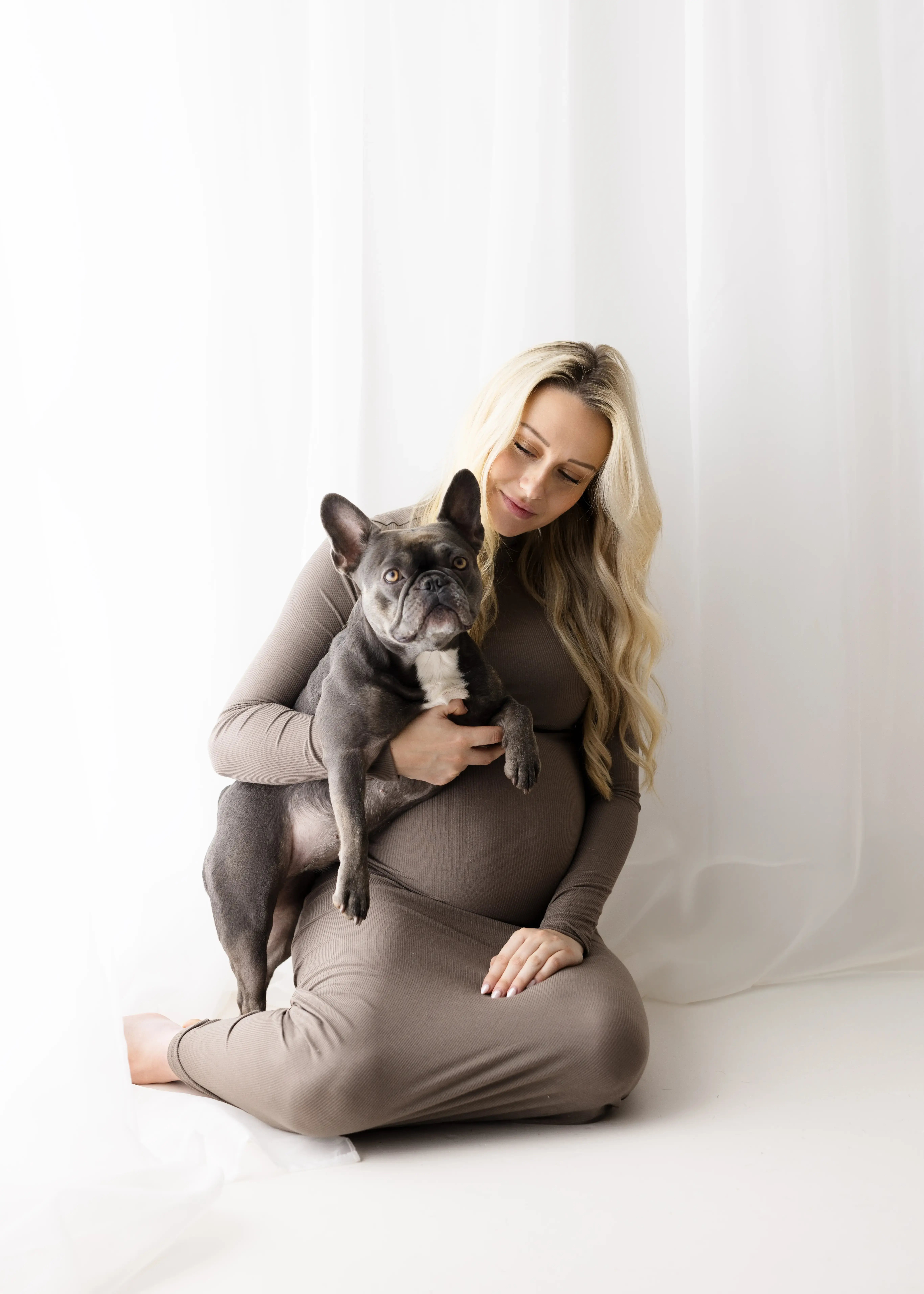 Expectant mother with long blonde hair wearing a snug brown dress, seated on the floor in a naturally lit studio, holding a grey French Bulldog on her lap.