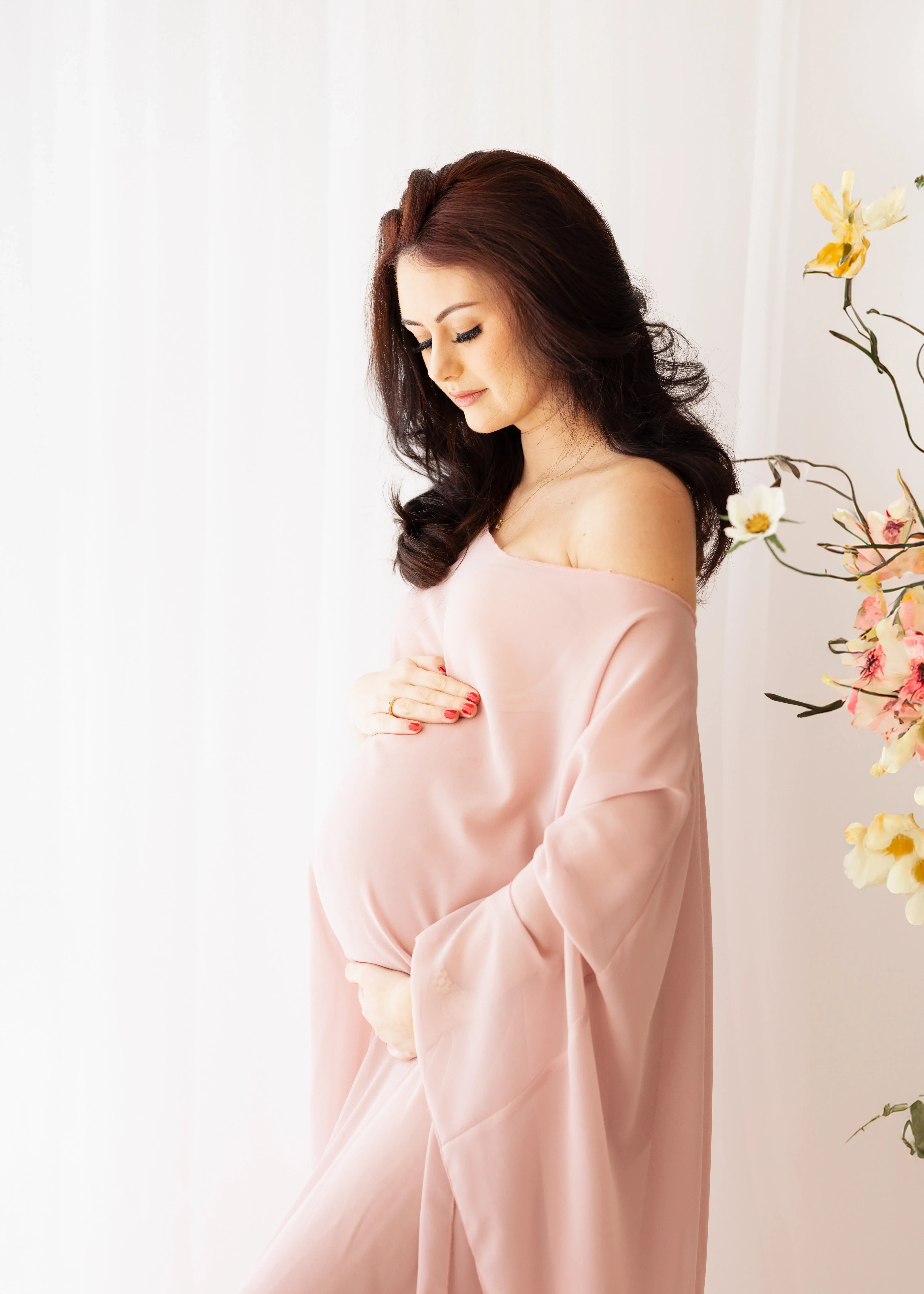 Side-profile of an expectant mother wearing a flowing pink drape, softly cradling her baby bump and gazing downward amid delicate spring flowers in a bright white studio.
