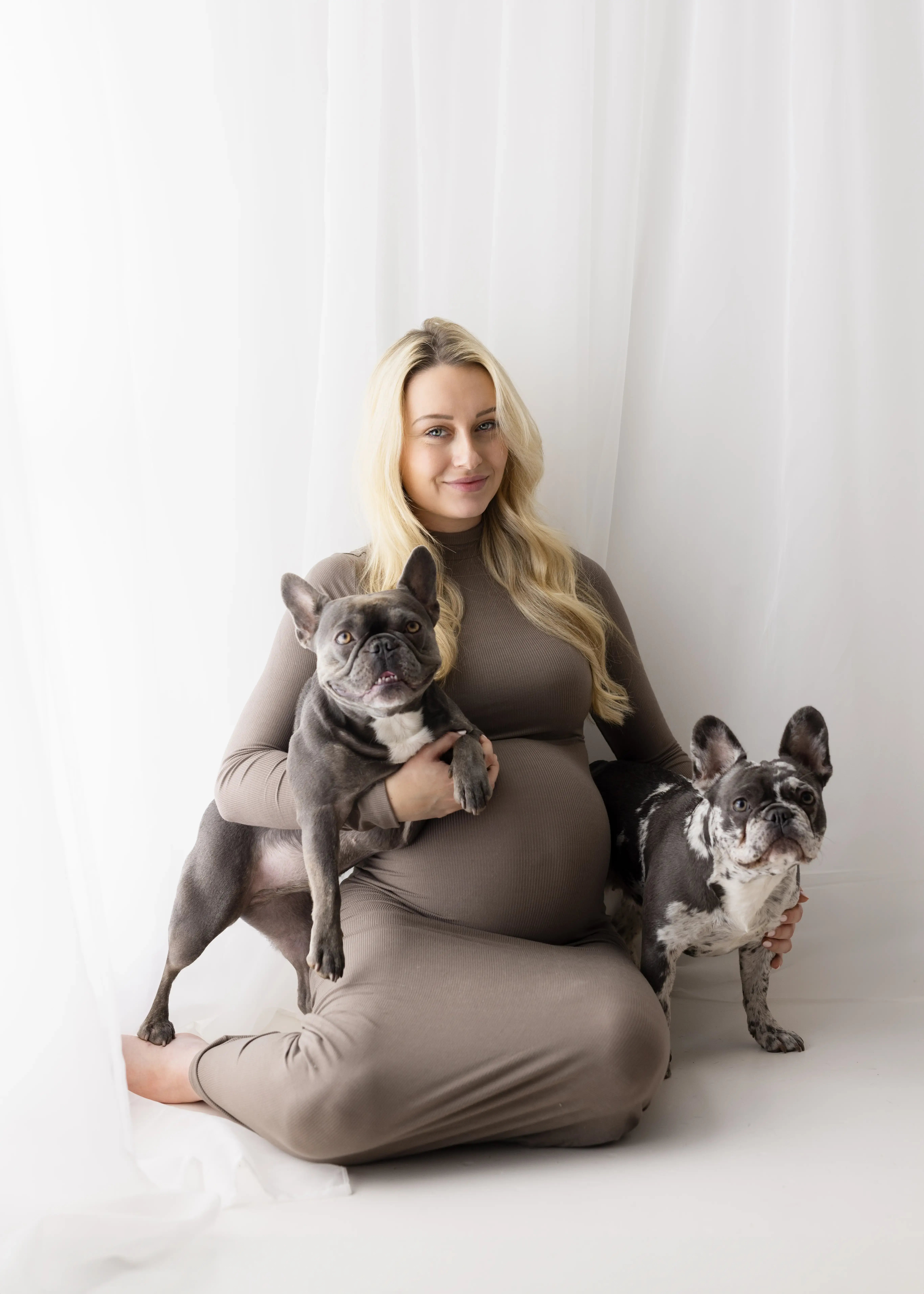 Smiling pregnant woman in a taupe maternity gown sitting in a white studio, lovingly accompanied by two French Bulldogs—one perched on her lap and the other by her side.