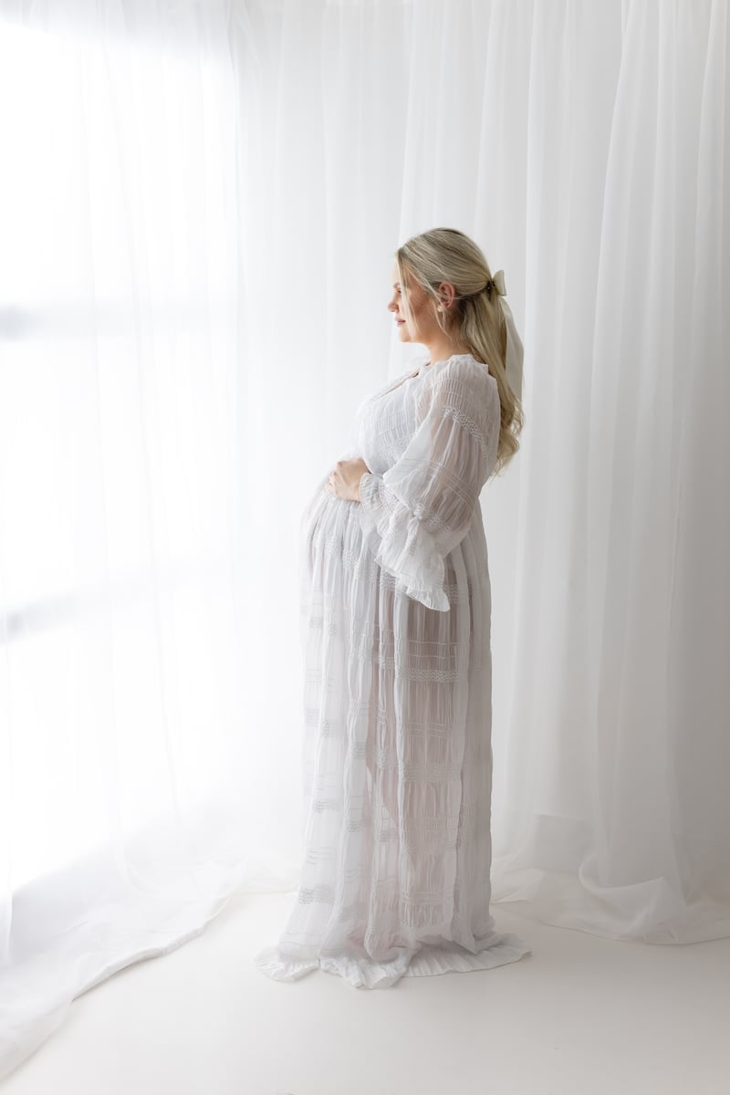 Mum-to-be looking out of a window in a white maternity gown for maternity photoshoot