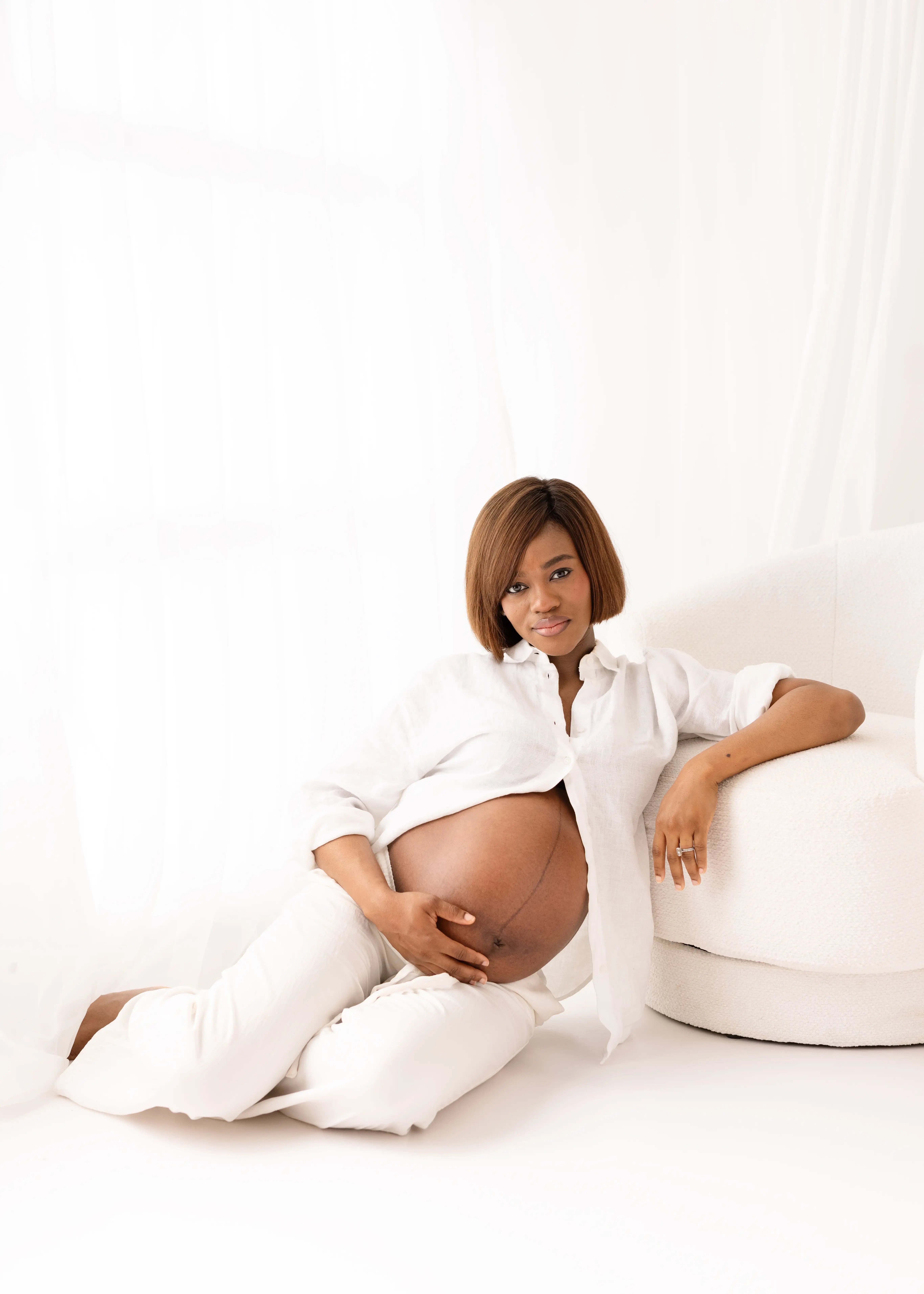 Pregnant woman dressed in white linen seated on the floor beside a white armchair, gently cradling her bare baby bump and looking calmly at the camera in a serene studio space.
