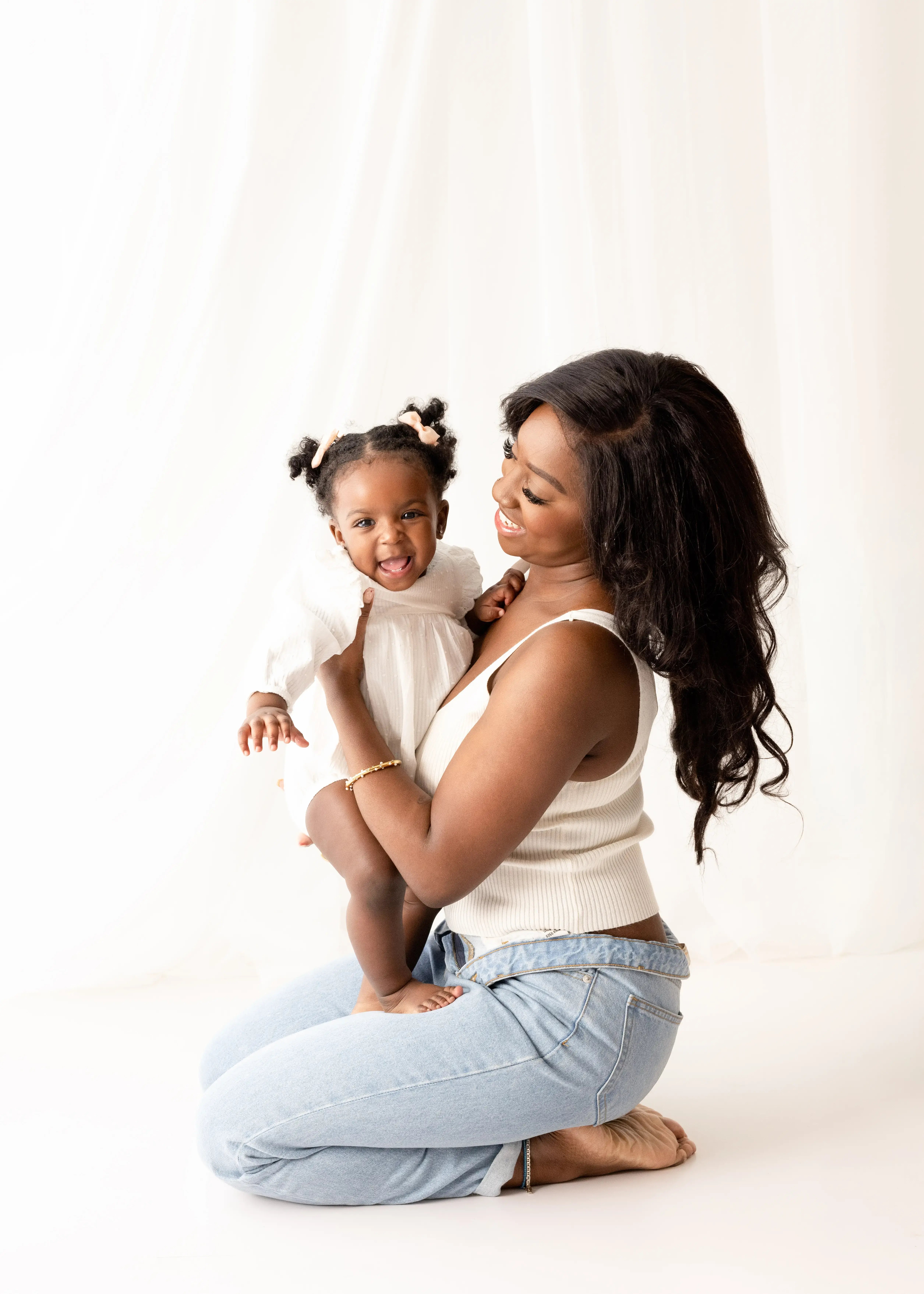 Smiling mother kneeling in light-wash jeans and a casual white top, holding her cheerful baby girl wearing a white dress and peach hair bows in a bright, airy mummy and me photoshoot.