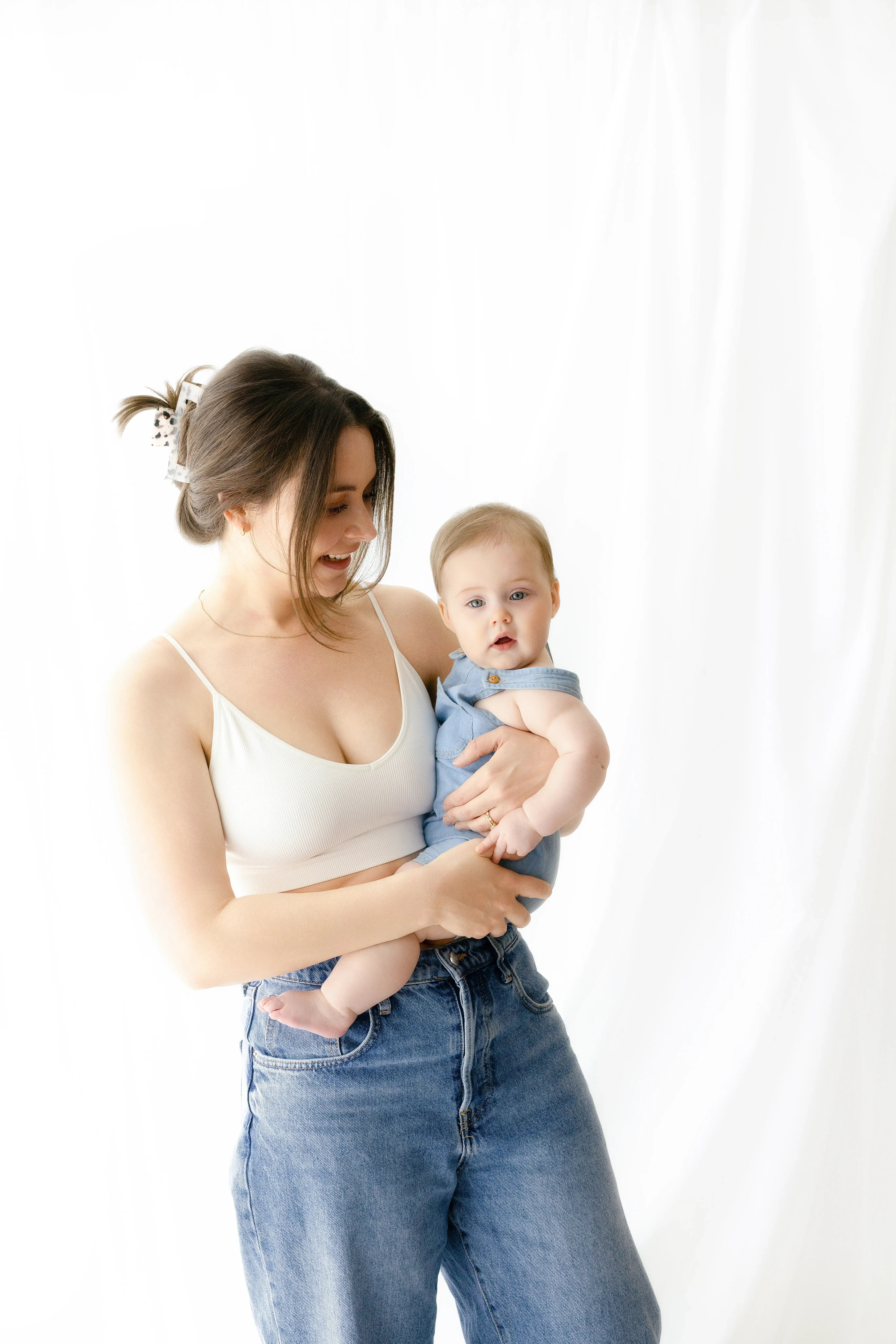 Mother wearing a simple white top and relaxed denim jeans holds her baby in a light denim playsuit, both smiling softly in front of a crisp white studio backdrop for a mummy and me portrait.