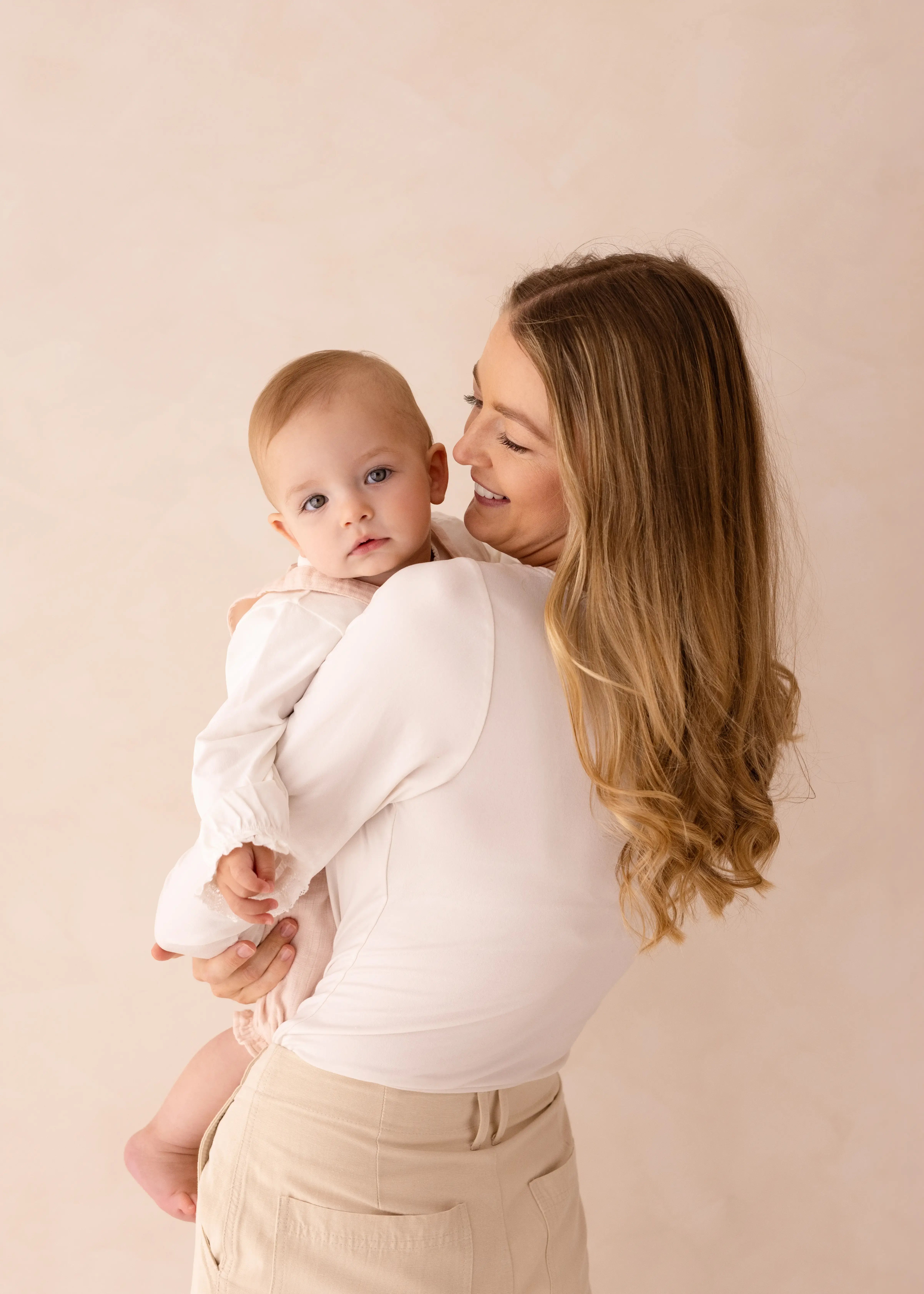A mother with long blonde hair smiles as she holds her baby girl close to her shoulder, both dressed in soft cream tones against a neutral backdrop.