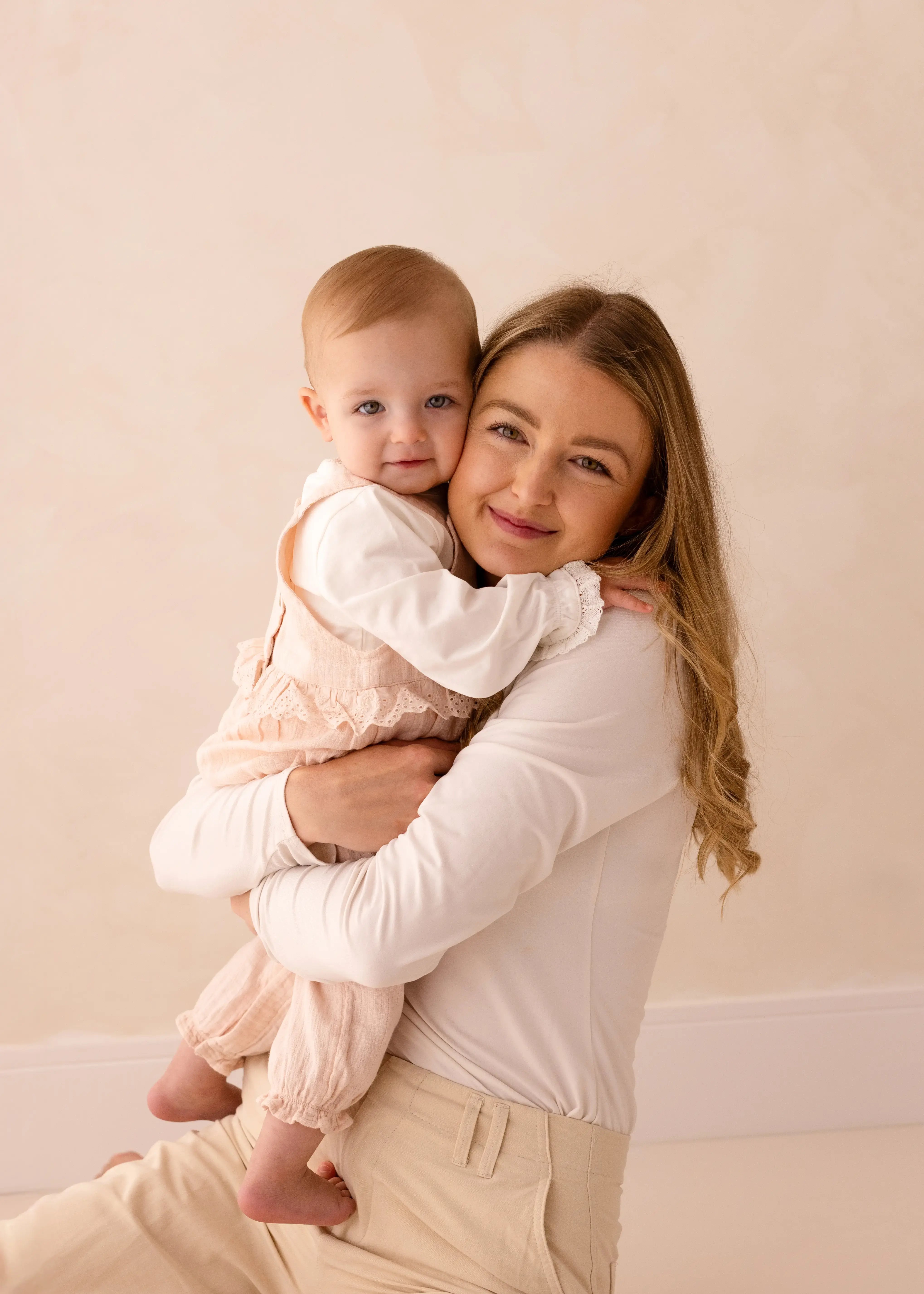 A mother kneels and hugs her baby girl closely, both smiling at the camera in matching neutral-toned outfits against a beige studio background.