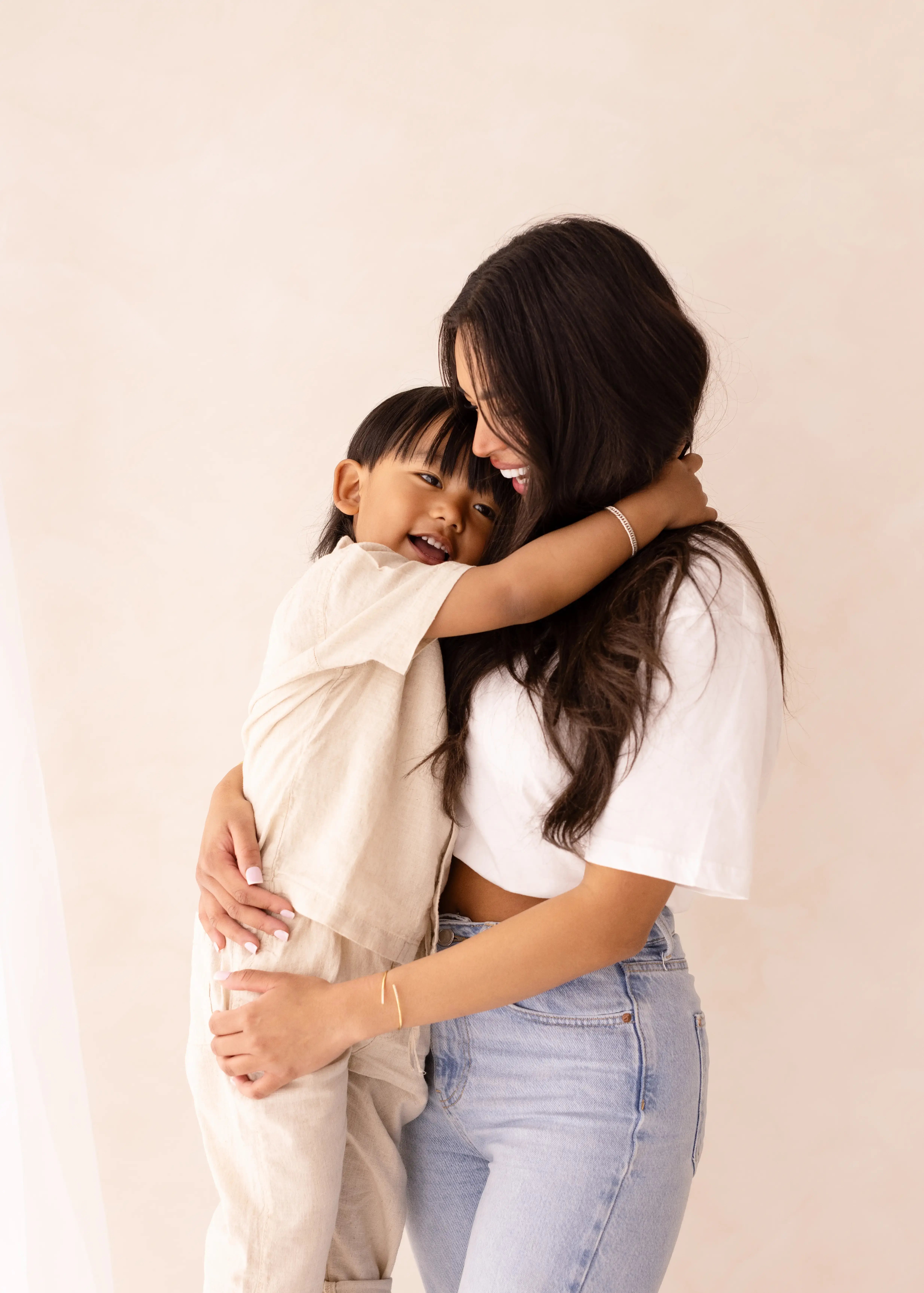 A mother holds her young child who wraps their arms around her neck, both smiling in matching cream and denim outfits against a soft neutral background.