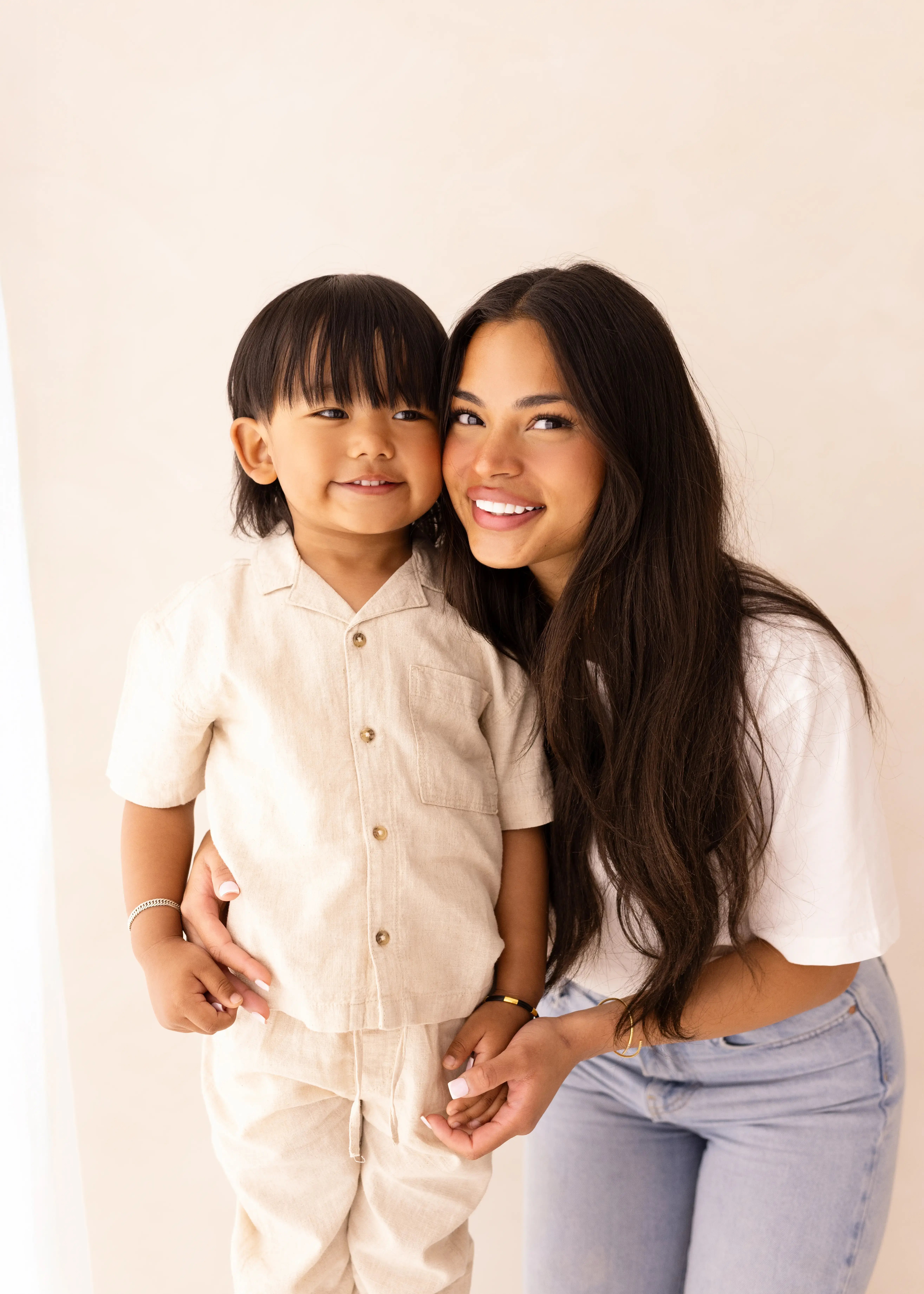 A mum and her toddler smile directly at the camera while standing close together in light-toned clothing, posed in front of a pale backdrop.