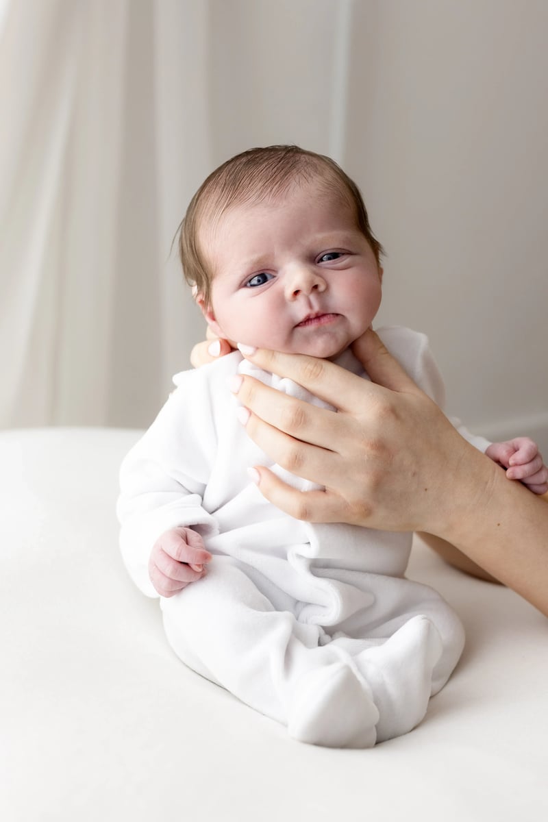 Awake newborn dressed in a cosy white sleepsuit, gently supported under the chin by a parent’s hand in a calm, light-filled portrait session.