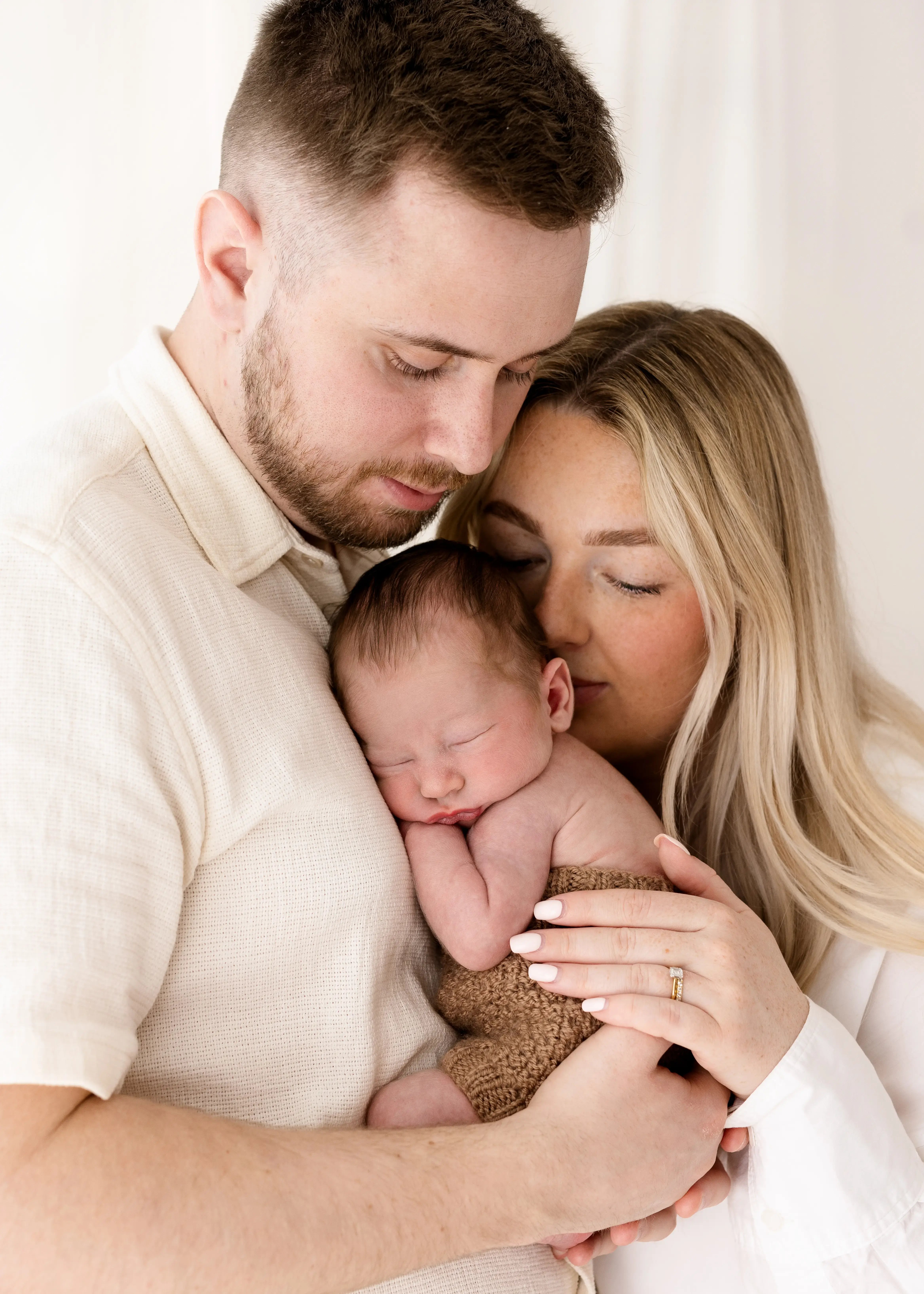 New parents holding their sleeping newborn baby close during a soft-lit family photoshoot.