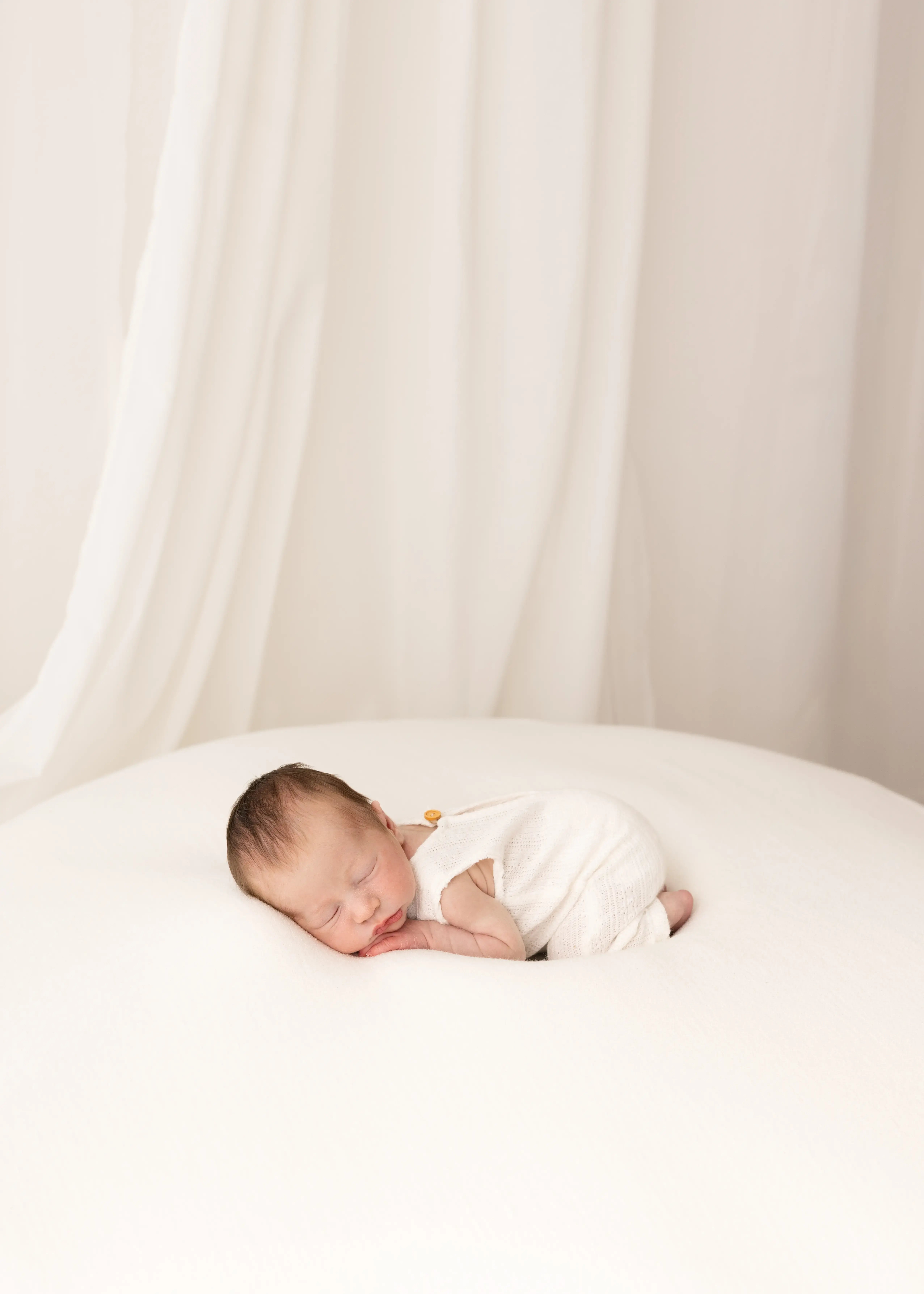 Newborn baby sleeping peacefully on a white blanket, wearing a white knit romper in a soft, minimal studio setting.