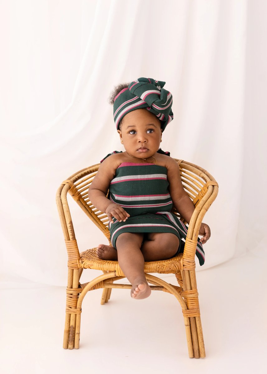 A baby girl wearing a green, pink, and white striped outfit and headwrap sits barefoot on a rattan chair with a neutral fabric backdrop behind her.