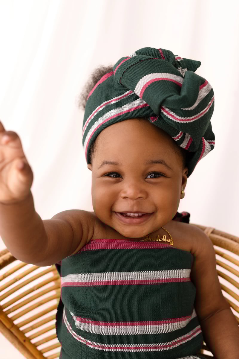 A happy baby girl in a green, pink, and white striped headwrap and dress reaches out with a wide smile, seated on a natural rattan chair in a softly lit studio.