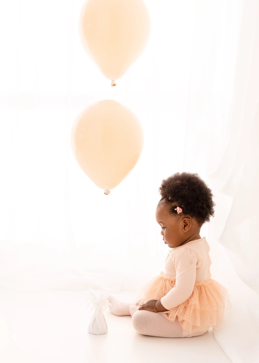 Toddler girl in a peach tutu looking at peach balloons during a milestone portrait session in a soft white studio.