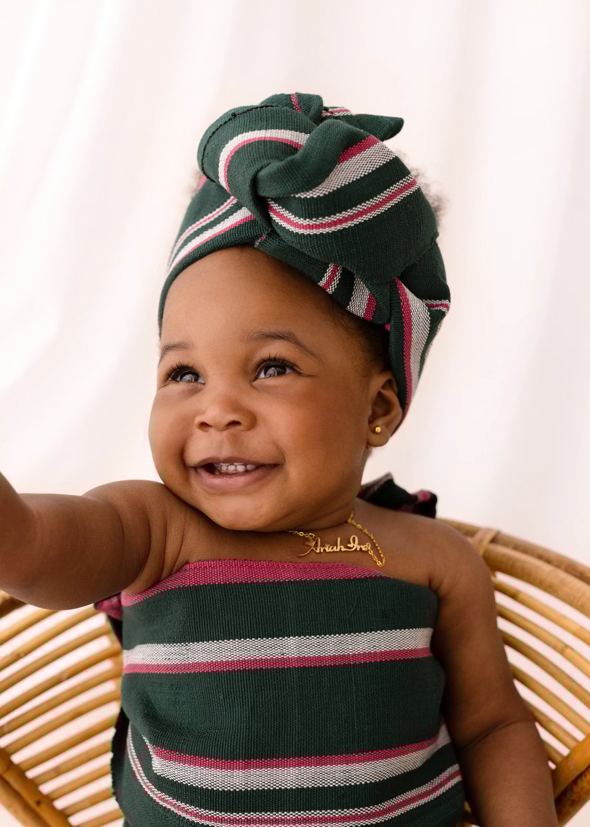 A baby girl smiles and looks upward while wearing a green, pink, and white striped headwrap and dress, seated in a wicker chair.